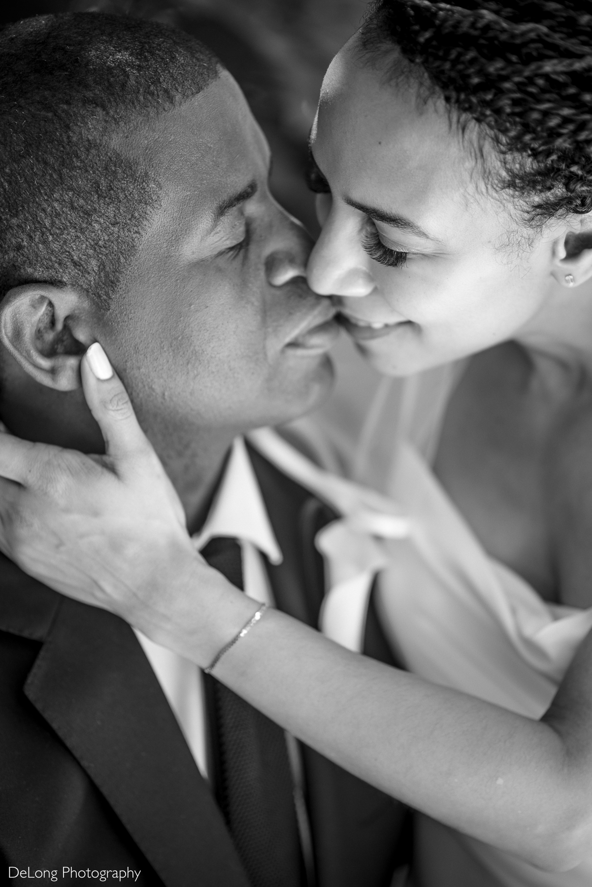 A black and white close-up photograph of a newlywed couple sharing an intimate moment in Uptown Charlotte, NC. The bride gently cradles the groom’s face as they lean in for a soft kiss, eyes closed and expressions full of love. The groom is dressed in a classic black suit and tie, while the bride’s delicate veil and gown add a touch of elegance. Captured by DeLong Photography.
