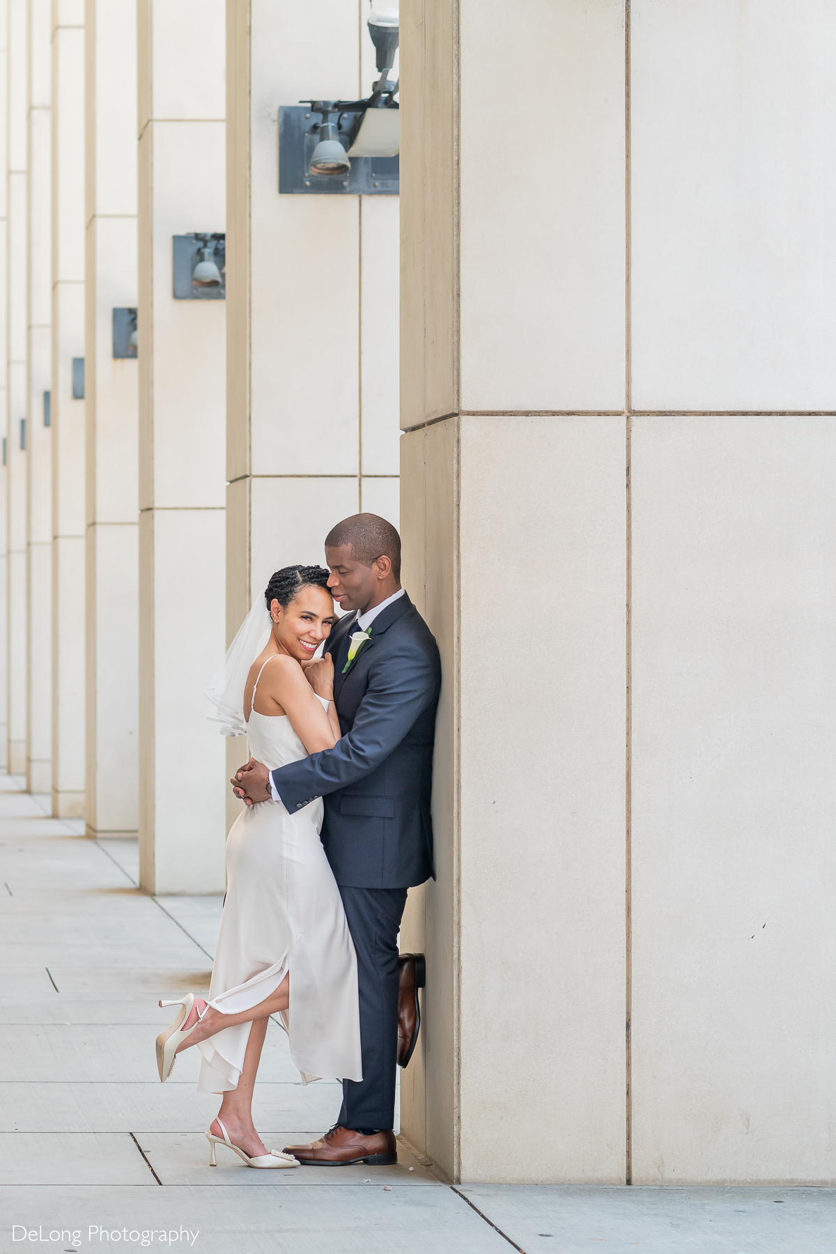 A romantic wedding portrait of a newlywed couple embracing against the modern architecture of the Charlotte Convention Center in Uptown Charlotte. The groom, dressed in a navy suit, leans against a pillar while holding his bride close. The bride, in a sleek white gown with a short veil, smiles radiantly as she leans into him. The elegant lines of the building and soft lighting create a timeless and intimate moment. Captured by DeLong Photography.