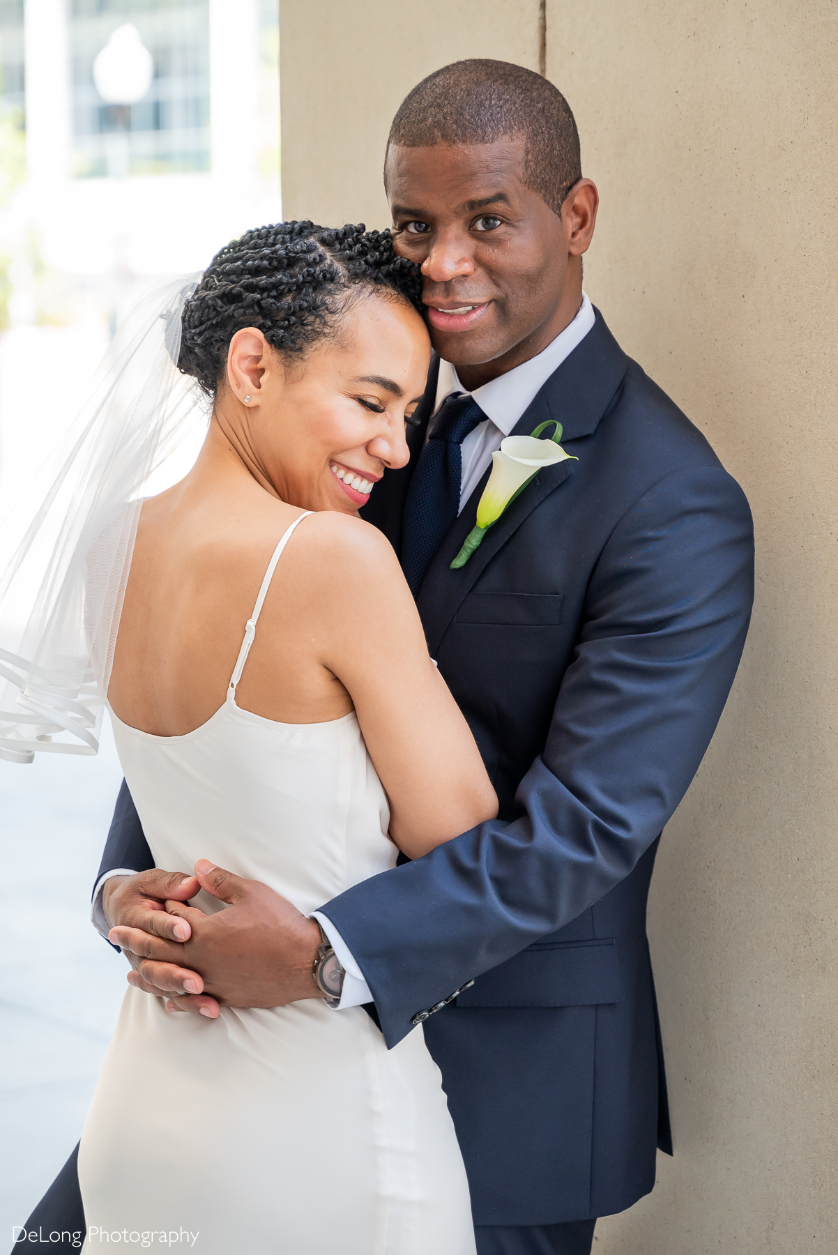 Elegant wedding portrait of a bride and groom embracing in Uptown Charlotte, NC. Bride in a minimalist white gown and veil, groom in a navy suit with calla lily boutonniere. Captured by DeLong Photography, Charlotte wedding photographers.