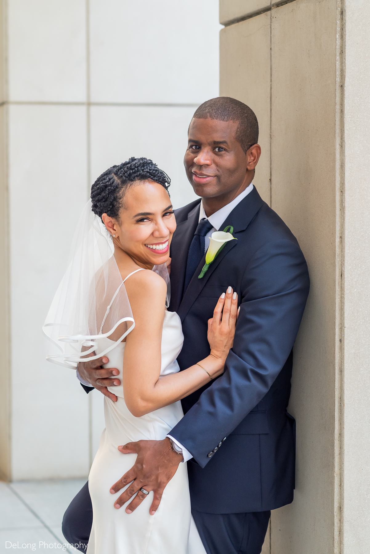 Modern wedding portrait of a bride and groom embracing in Uptown Charlotte, NC. The bride wears a sleek white gown and veil, smiling brightly, while the groom stands in a navy suit with a white calla lily boutonniere. Captured by DeLong Photography, Charlotte wedding photographers specializing in urban and intimate weddings.
