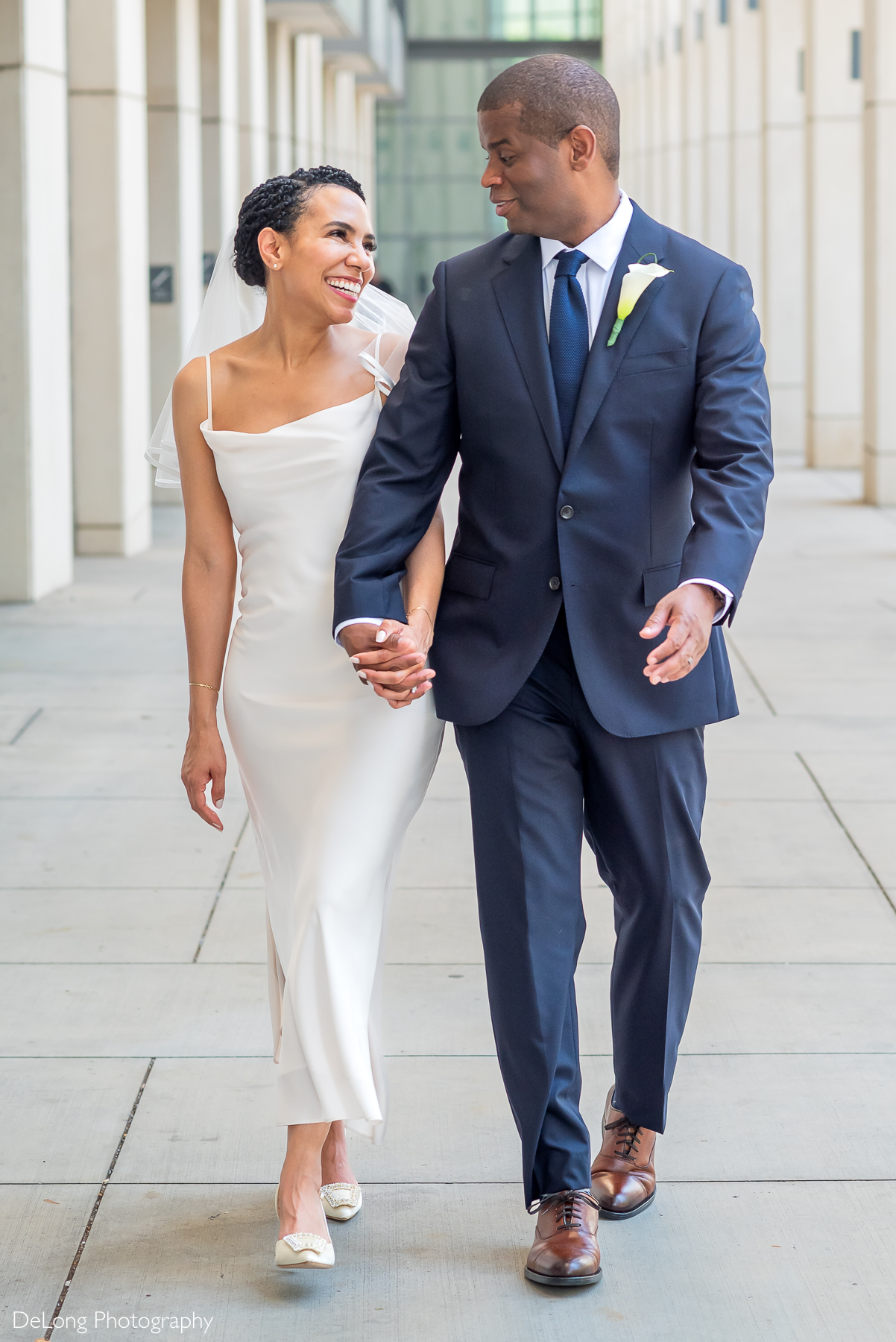 Joyful bride and groom walking hand-in-hand through Uptown Charlotte during their stylish elopement. Bride wears a modern white gown and flats, groom in a navy suit with calla lily boutonniere. Captured by DeLong Photography, Charlotte elopement photographers specializing in intimate urban weddings.