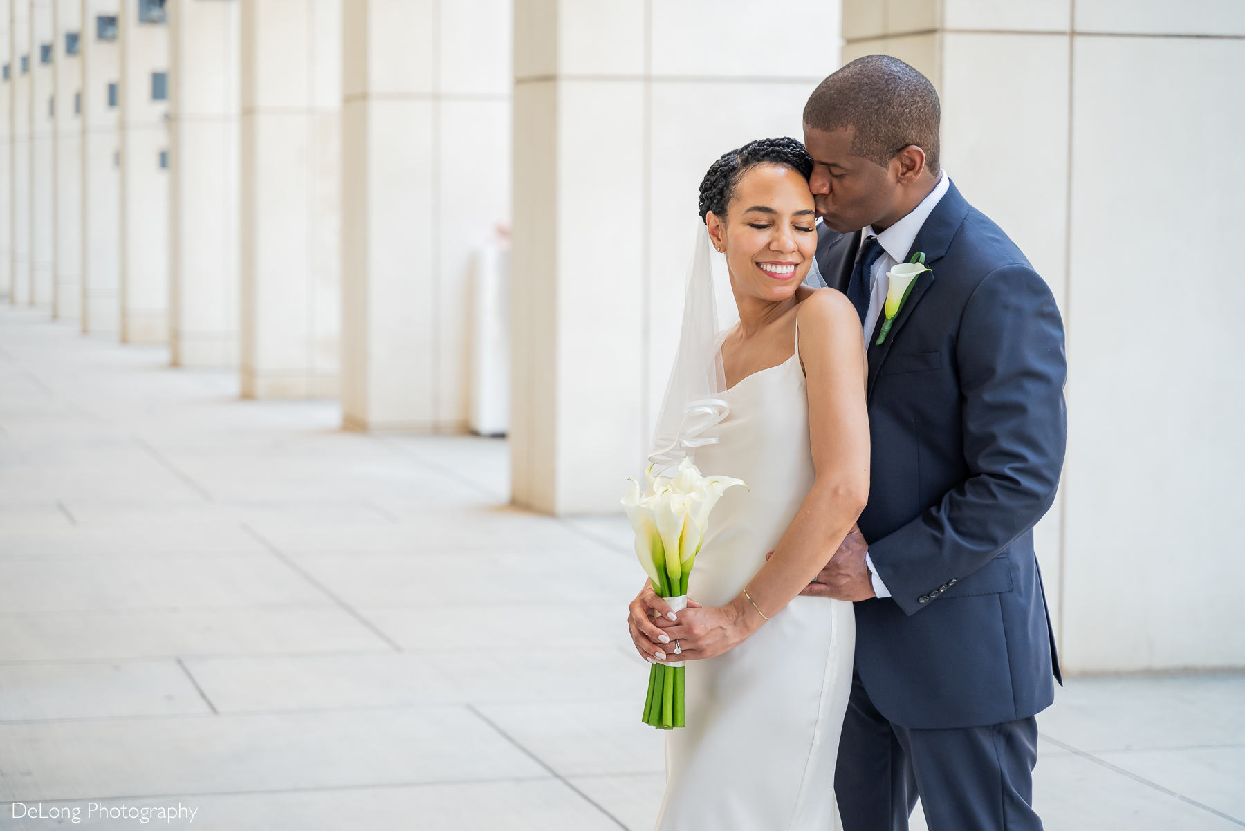 Groom kissing bride’s temple as she smiles, holding white calla lilies in Uptown Charlotte. Captured by DeLong Photography, Charlotte NC elopement photographers specializing in intimate wedding portraits.