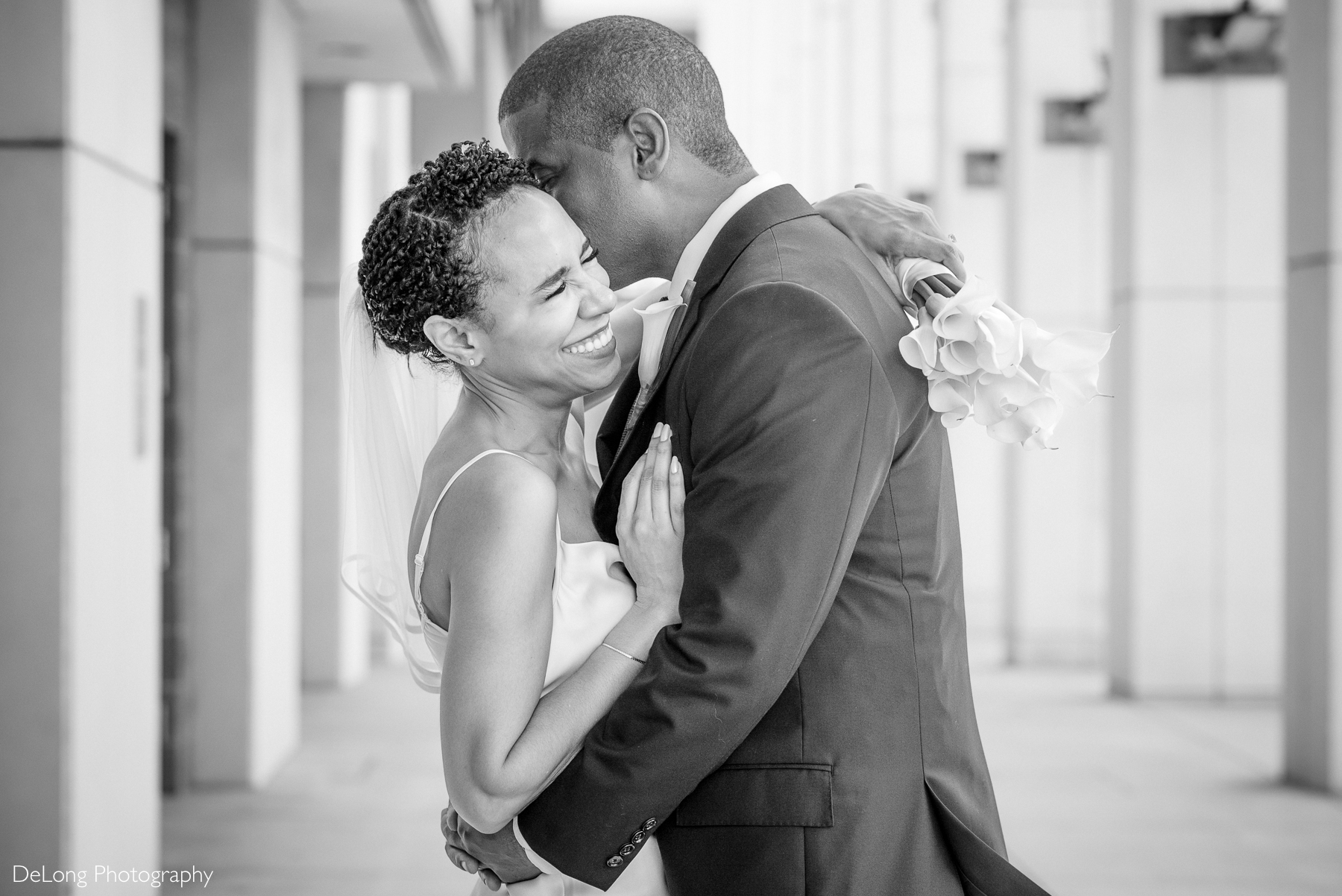 Joyful bride laughing as groom embraces her and holds calla lilies in Uptown Charlotte. A candid moment captured by DeLong Photography, Charlotte elopement photographers specializing in intimate, modern wedding portraits.