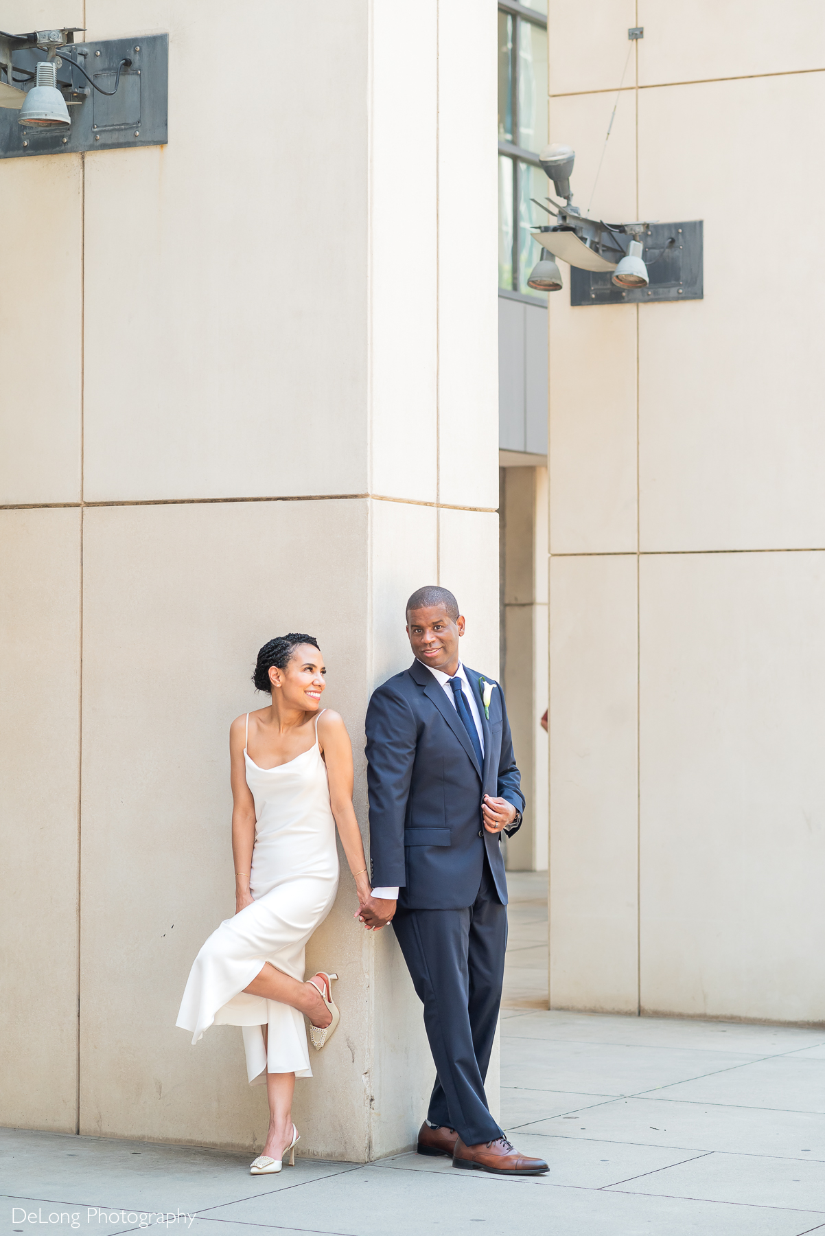A stylish bride and groom share a quiet moment around the corner of a modern architectural column in Uptown Charlotte. The bride, in a sleek ivory slip dress and short veil, smiles brightly at her groom while holding his hand, her heel kicked up playfully. The groom wears a tailored navy suit with a calla lily boutonniere, gazing at her with a subtle smile. Captured by DeLong Photography, this elegant portrait blends urban sophistication with heartfelt connection—perfect inspiration for engaged couples seeking a luxury wedding photographer in Charlotte, NC.