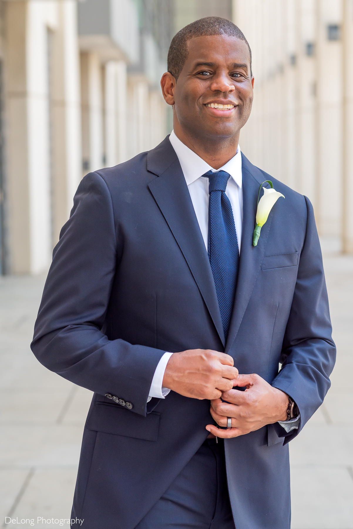 Groom in a tailored navy suit with calla lily boutonniere standing confidently in Uptown Charlotte, captured by DeLong Photography—Charlotte wedding photographers known for timeless, luxury portraits with a modern editorial style.