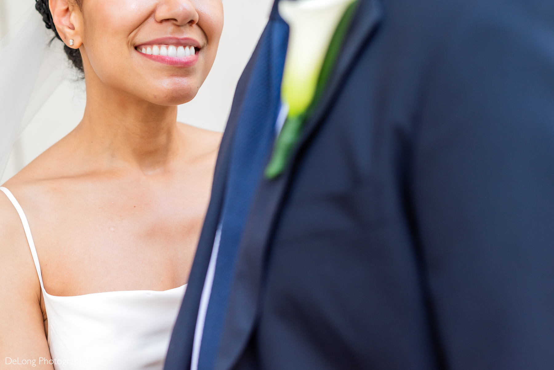 Close-up of a smiling bride in a minimalist satin gown during a modern wedding in Uptown Charlotte, with the groom in a navy suit and calla lily boutonniere in the foreground. Captured by DeLong Photography, a luxury Charlotte wedding photographer specializing in elegant and editorial-style wedding portraits.