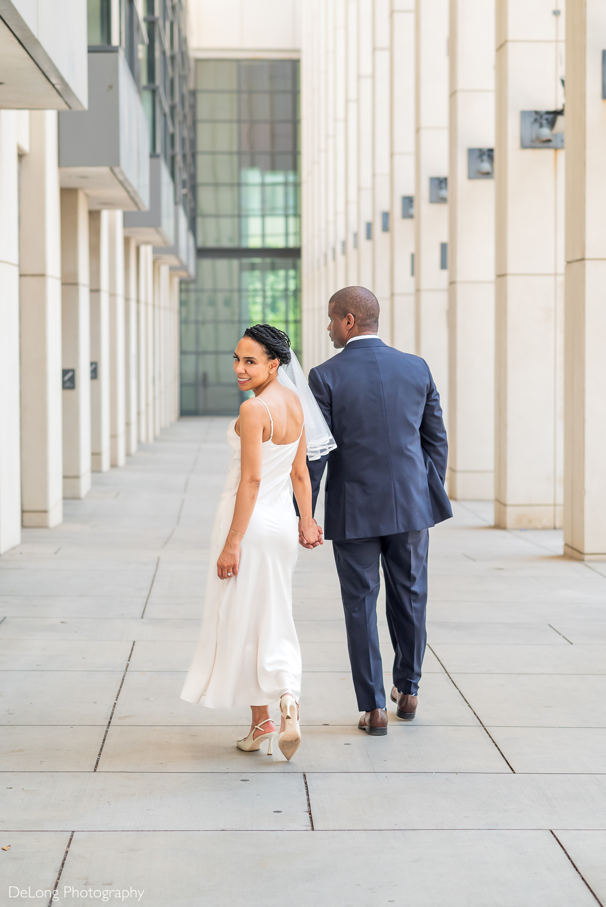 A stylish bride in a satin gown and veil walks hand-in-hand with her groom in a navy suit through the modern architecture of Uptown Charlotte. Captured by DeLong Photography, a top luxury wedding photographer in Charlotte, NC, known for elegant, editorial-style wedding portraits in urban settings.