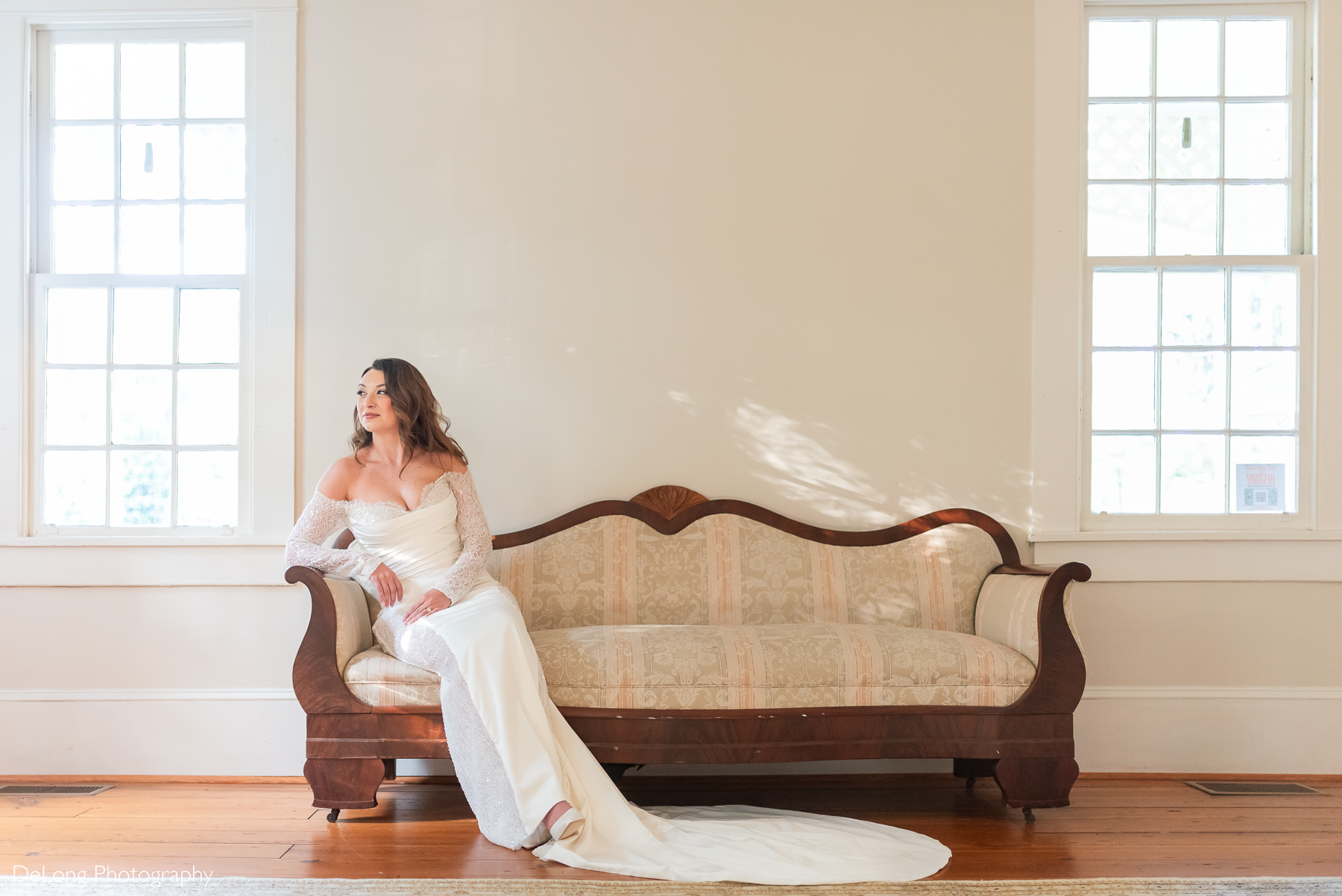 Carrie poses elegantly on a vintage sofa beneath soft natural light during her bridal portrait session at The White Home in Rock Hill, SC, wearing an off-the-shoulder gown with beaded lace sleeves. Photographed by DeLong Photography.