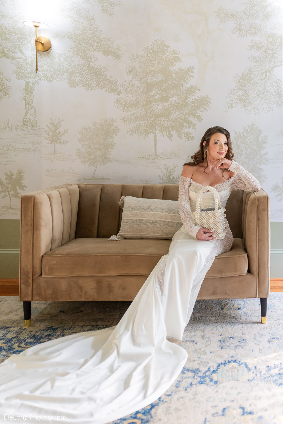 Bridal portrait at The White Home in Rock Hill, SC, featuring a bride in an off-the-shoulder lace wedding gown with a long train, seated on a velvet couch in front of elegant toile wallpaper, captured by DeLong Photography.