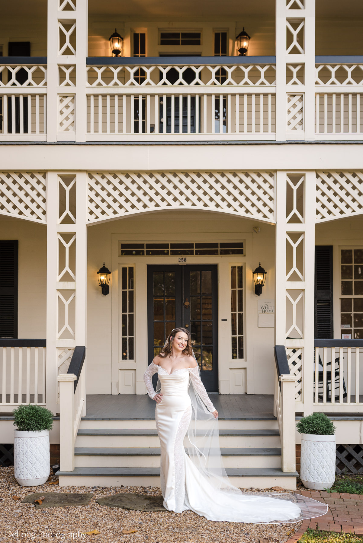 Full-length bridal portrait in front of the historic entrance of The White Home in Rock Hill, SC, featuring the bride in a fitted gown with sheer sleeves and a long veil, taken by DeLong Photography.