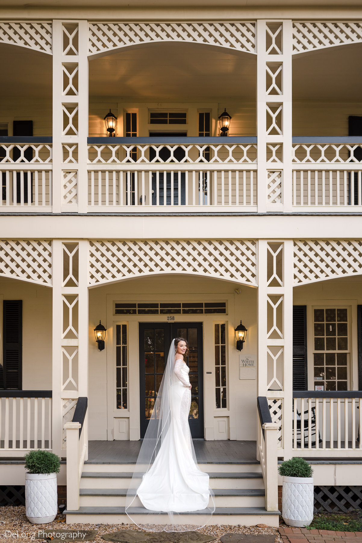 Elegant bridal portrait on the front steps of The White Home in Rock Hill, SC. The bride wears a long-sleeved wedding gown with a dramatic train and veil, captured by DeLong Photography.
