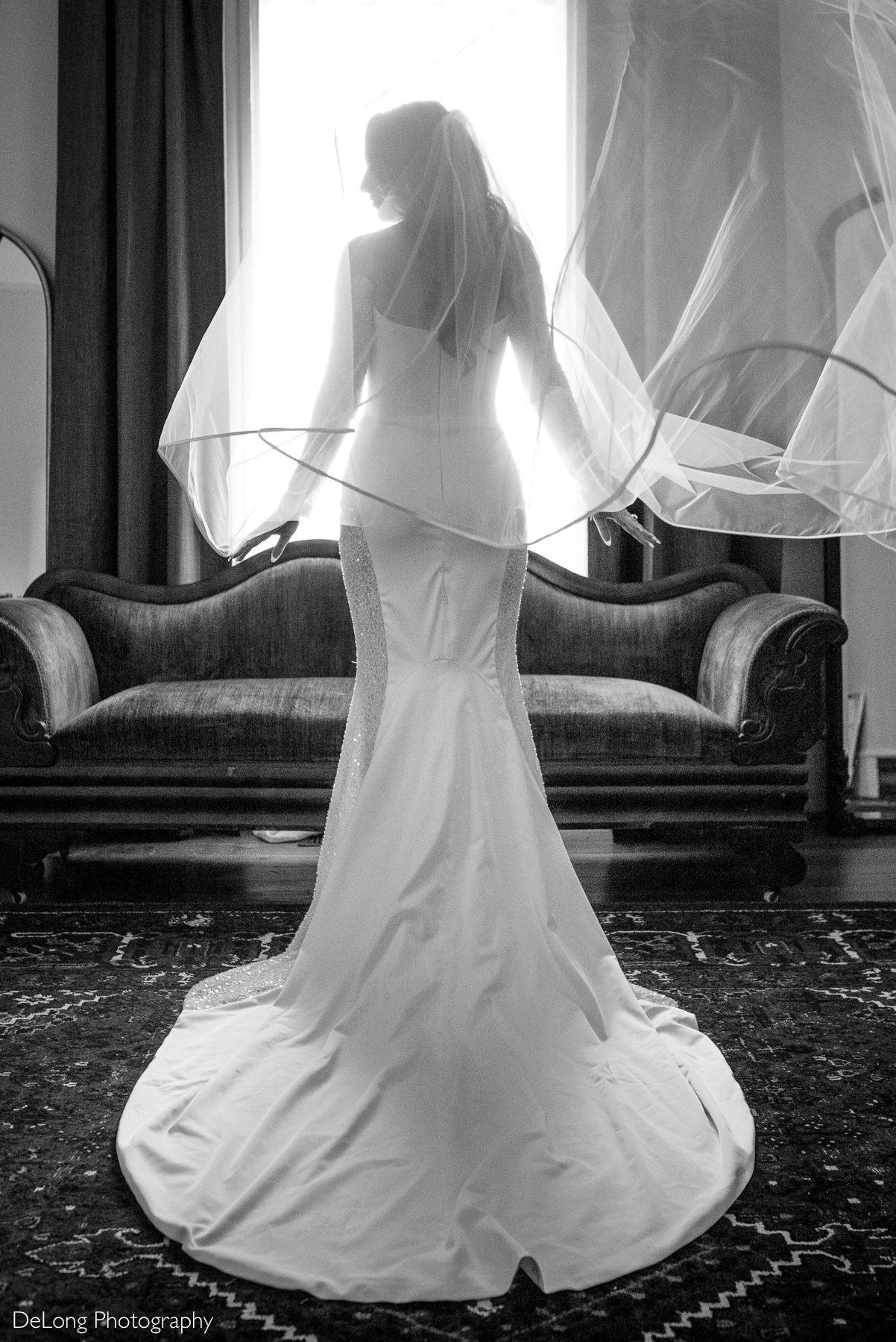 Bridal portrait of a bride standing in front of a window at The White Home in Rock Hill, SC, wearing a fitted wedding gown with a long train and veil flowing in motion. Captured by DeLong Photography.