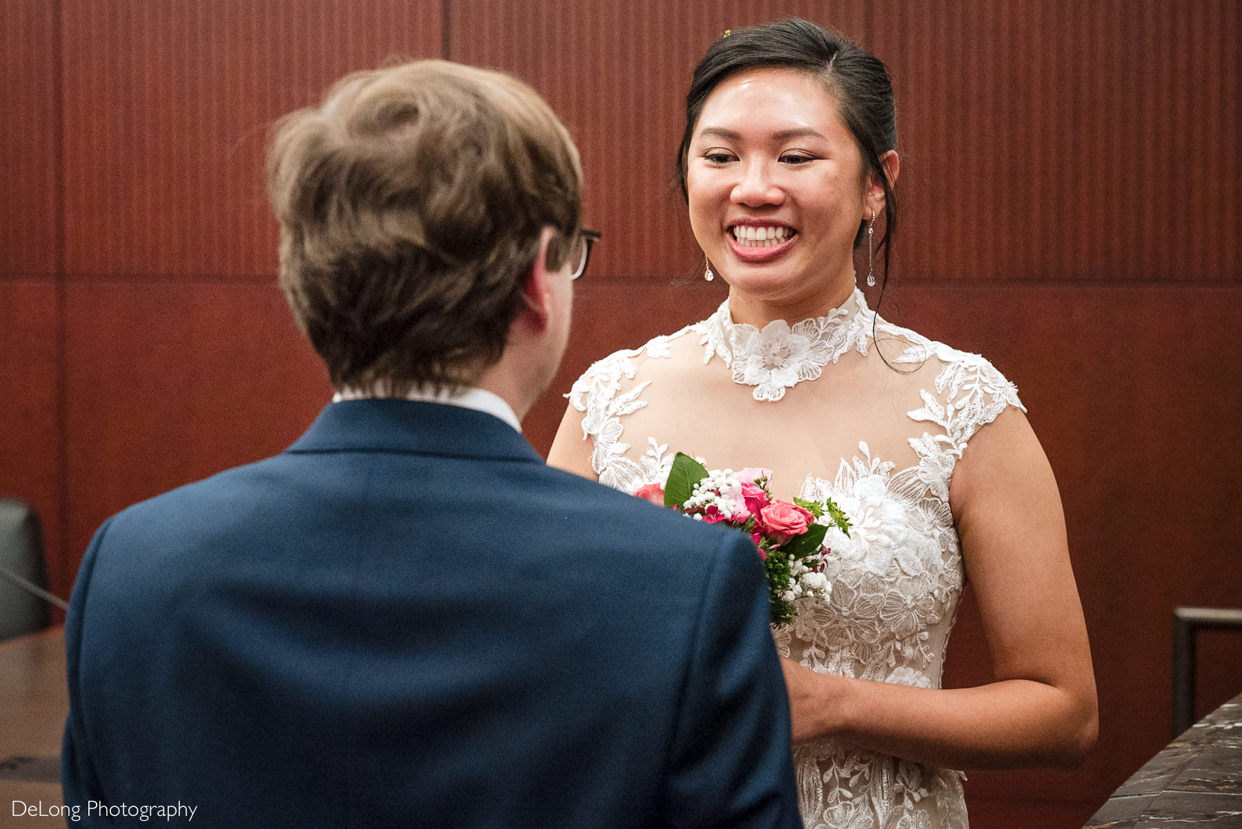 Bride smiles joyfully at her groom during their Raleigh elopement ceremony at the Wake County Courthouse, wearing a lace wedding dress and holding a pink bouquet — captured by DeLong Photography.
