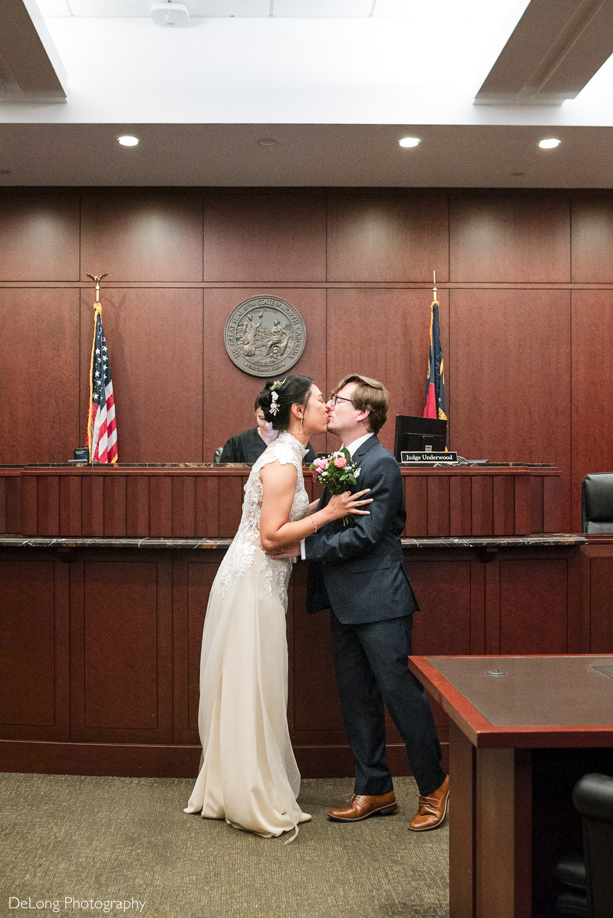 Bride and groom share their first kiss in the Wake County Courthouse courtroom during their Raleigh elopement ceremony, with Judge Underwood officiating — photographed by DeLong Photography.
