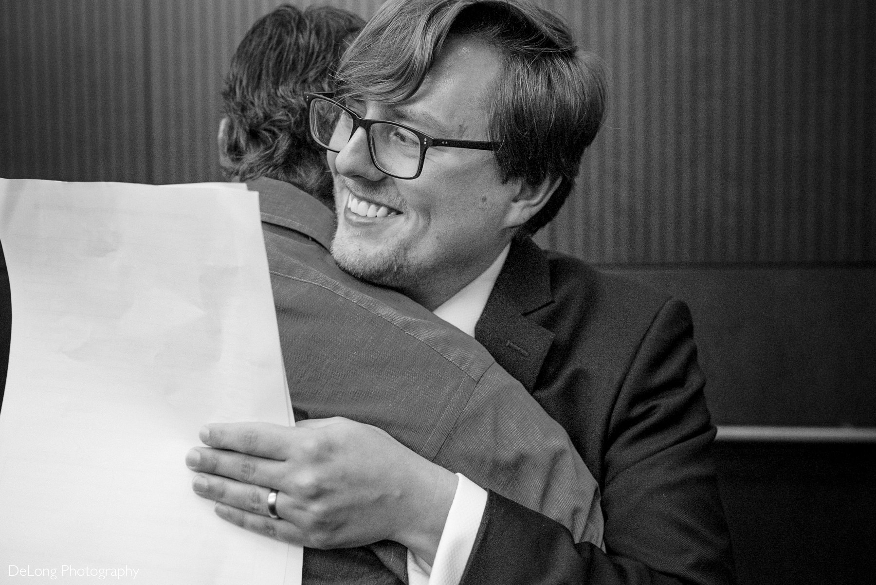 Groom smiles and hugs a loved one while holding marriage paperwork during a joyful moment at his Raleigh elopement at the Wake County Courthouse — captured by DeLong Photography.