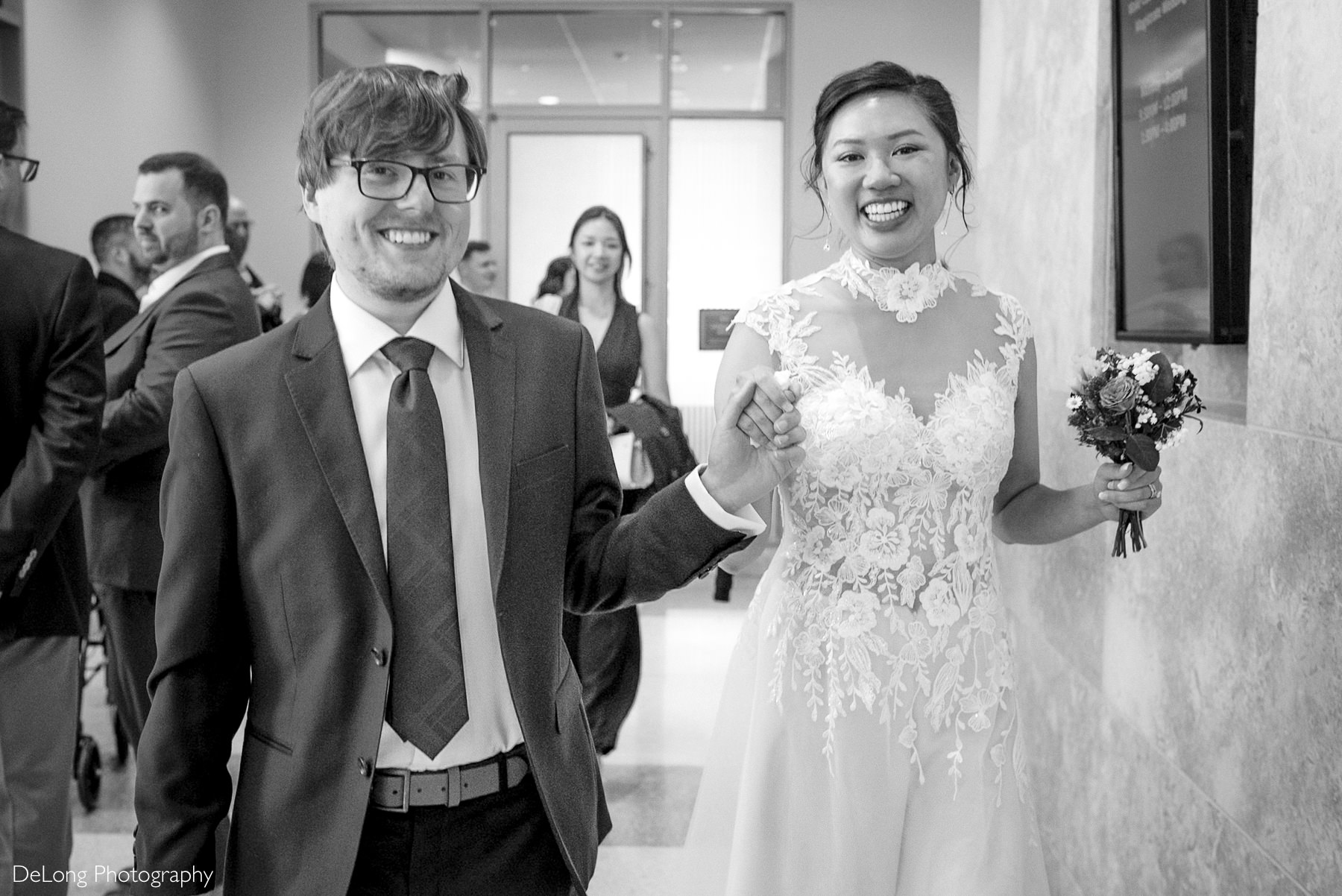 Just married couple walks hand in hand through Wake County Courthouse during their Raleigh elopement — bride smiling in a floral lace gown, groom in a dark suit, surrounded by joyful guests. Captured by DeLong Photography.