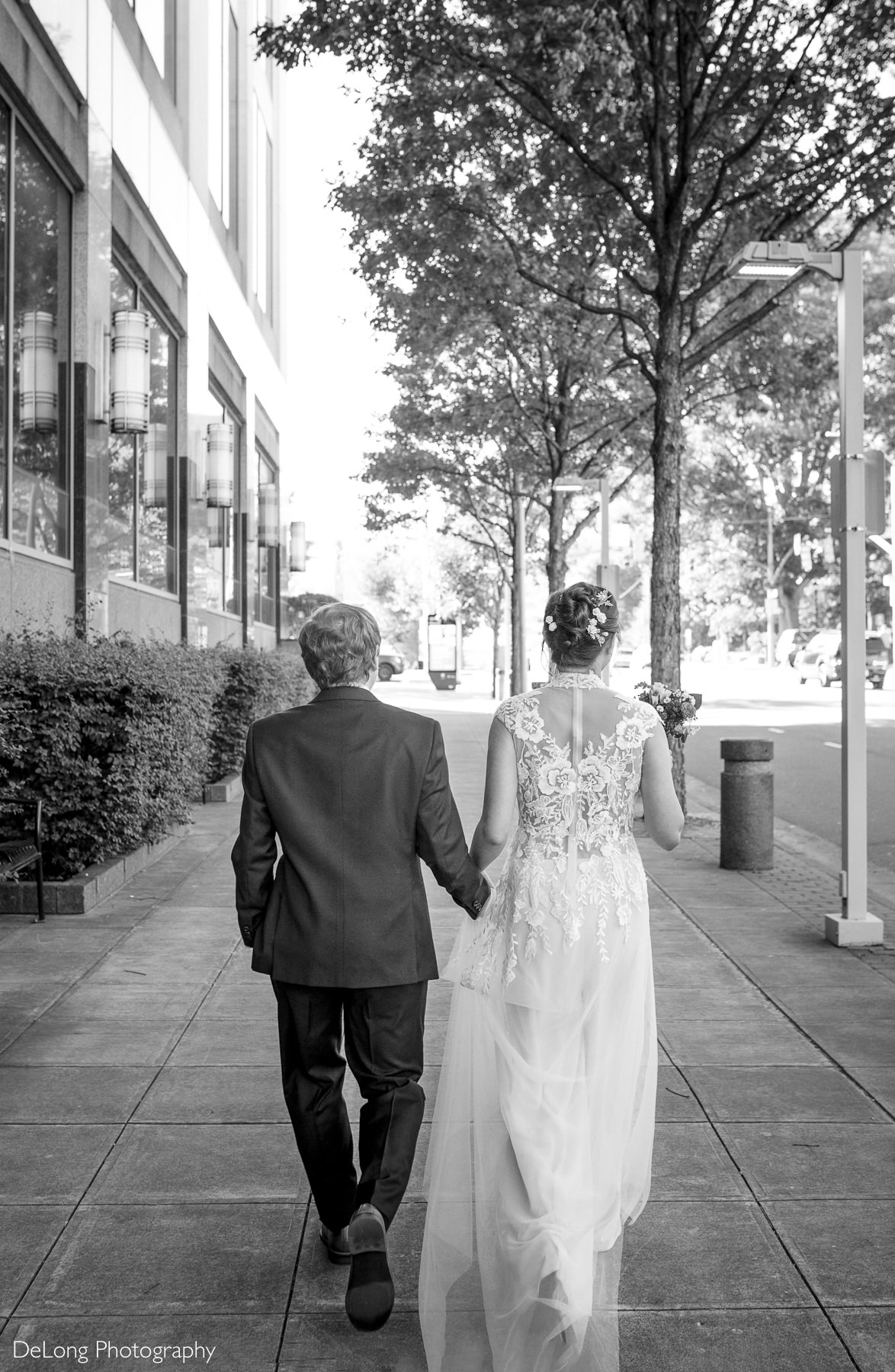 Bride and groom walk hand-in-hand down a downtown Raleigh sidewalk after their Wake County Courthouse elopement. The bride’s lace wedding dress flows behind her as the couple strolls past modern buildings and tree-lined streets. Captured by DeLong Photography.