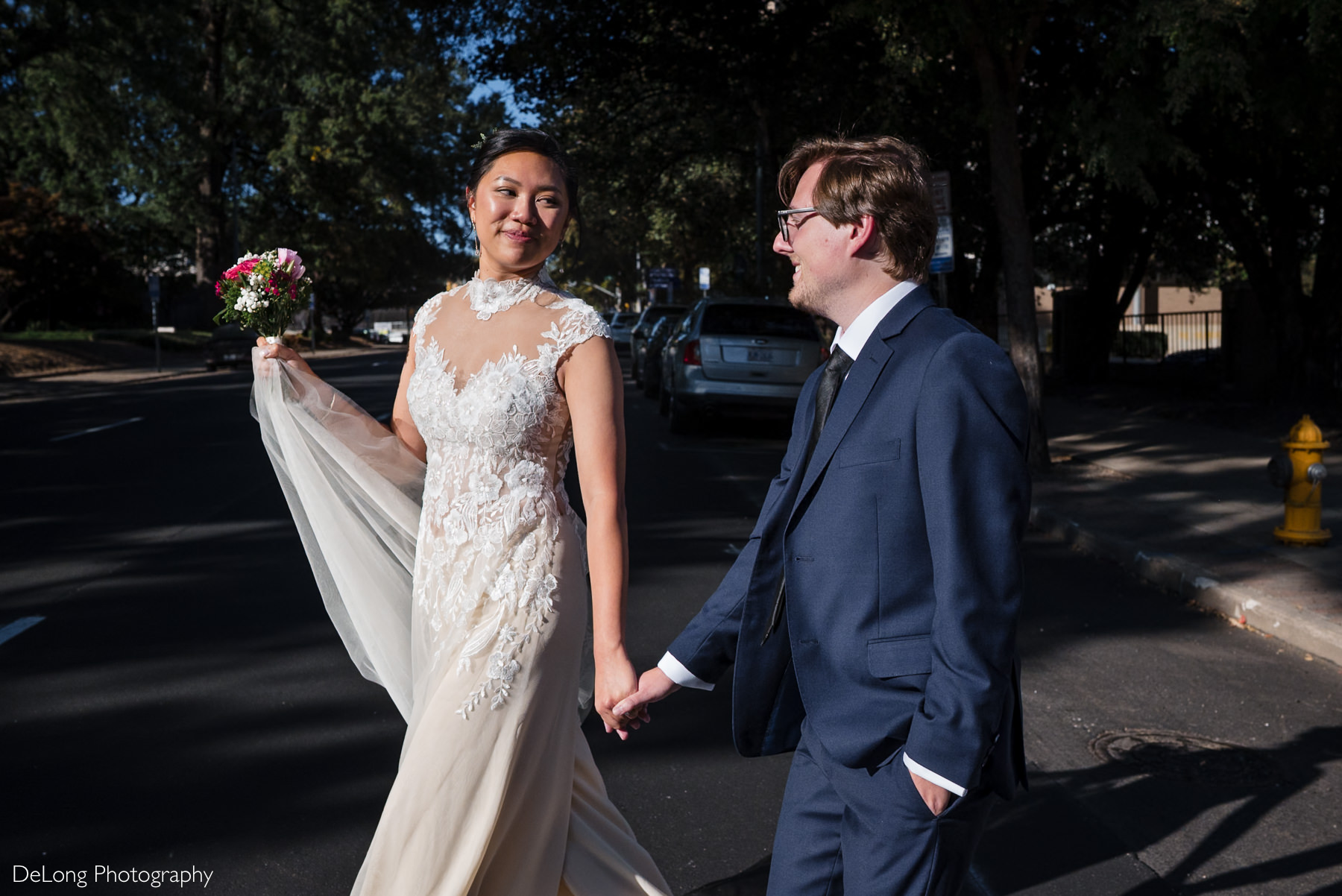 Bride and groom hold hands while crossing the street during their Wake County Courthouse elopement in downtown Raleigh, NC. The bride wears a lace high-neck wedding gown and holds a bouquet, while the groom wears a navy suit. Captured by DeLong Photography.