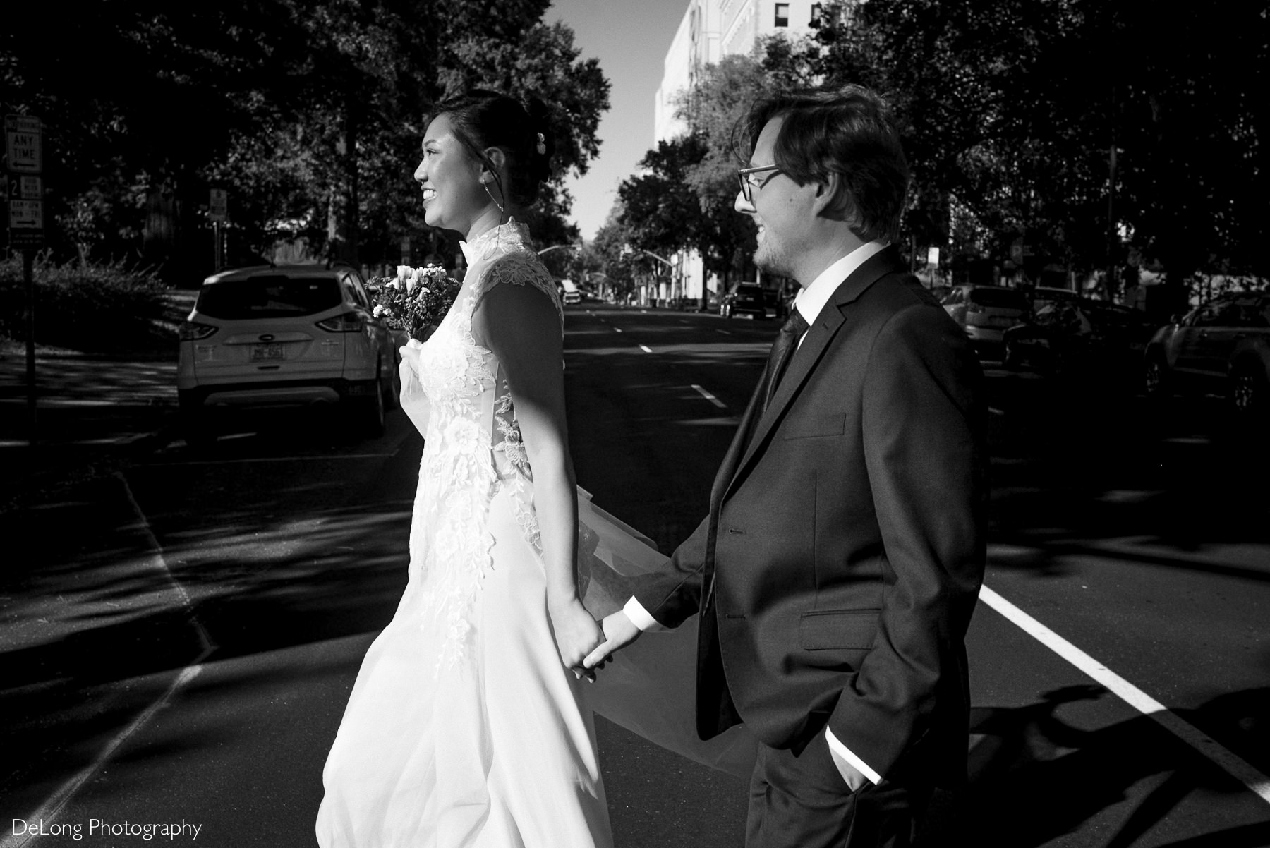 Black and white photo of a joyful bride and groom holding hands while crossing the street during their downtown Raleigh elopement near the Wake County Courthouse. The bride wears a lace wedding gown with floral details and carries a bouquet. Captured by DeLong Photography.