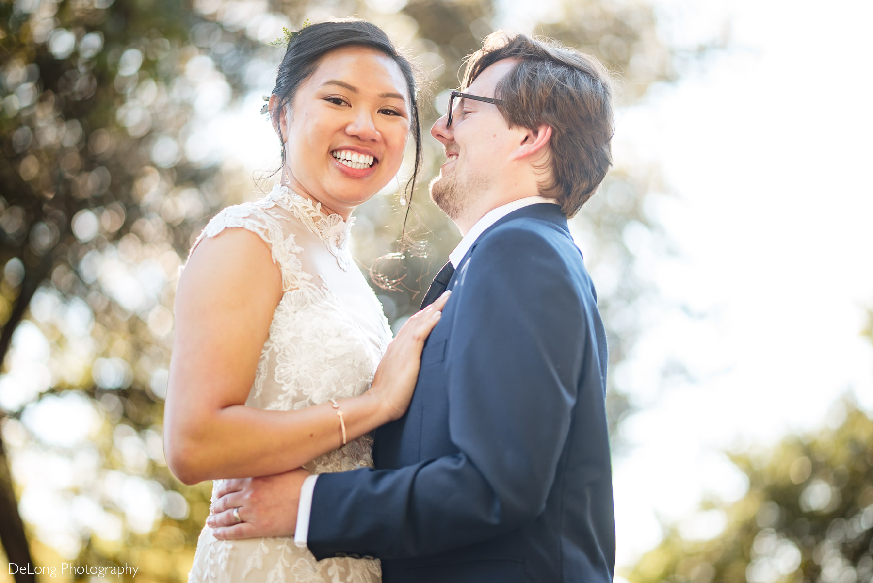 Bride and groom smile joyfully during their intimate Wake County Courthouse elopement in Raleigh, North Carolina. The bride wears a lace high-neck wedding gown, while the groom is in a classic navy suit. Captured by DeLong Photography under glowing sunlight.
