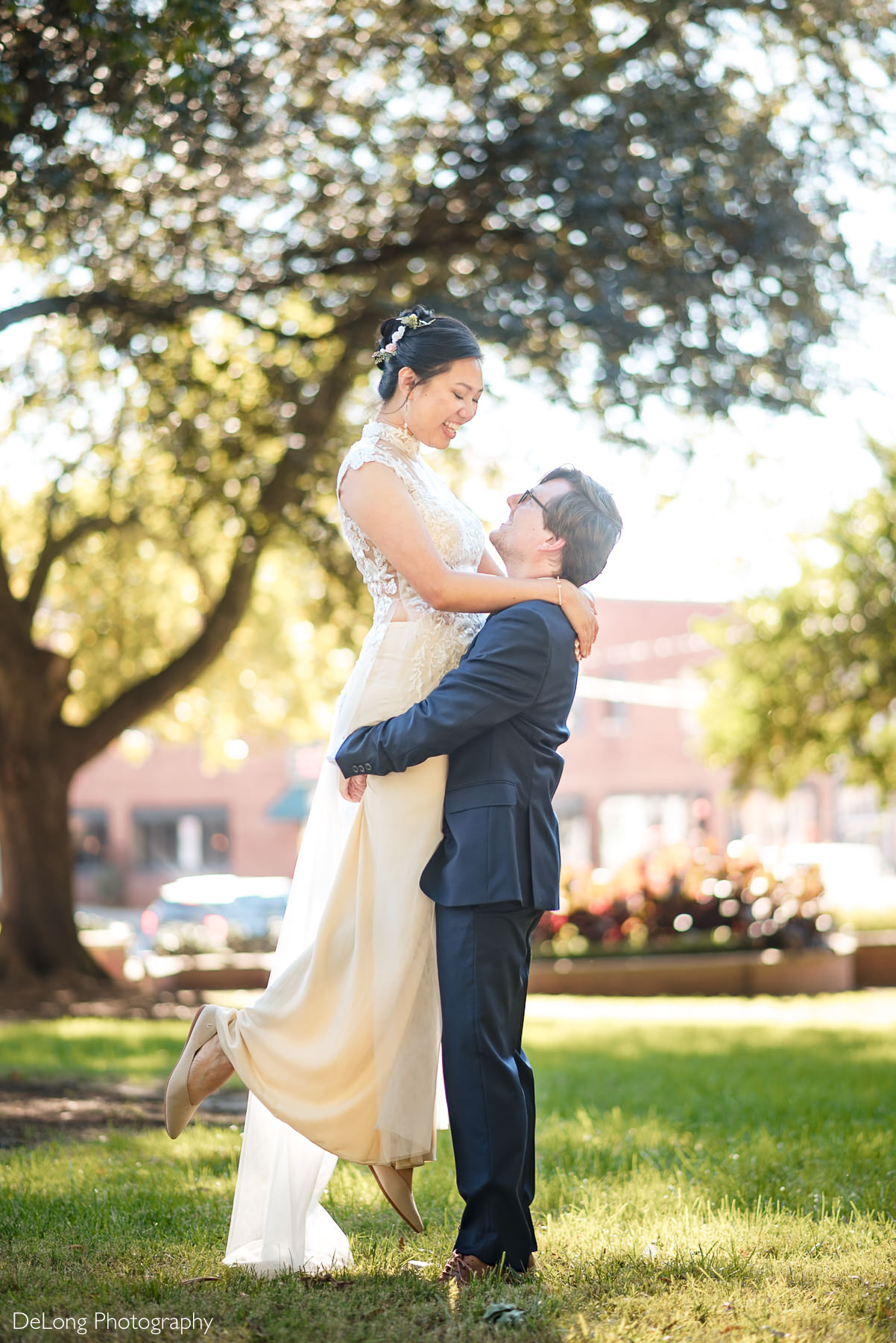 Groom lifts bride in a joyful embrace during their Raleigh elopement at sunlit Nash Park near the Wake County Courthouse. The bride wears a sleeveless lace gown with floral hair accessories, and the groom wears a navy suit. Captured by DeLong Photography.