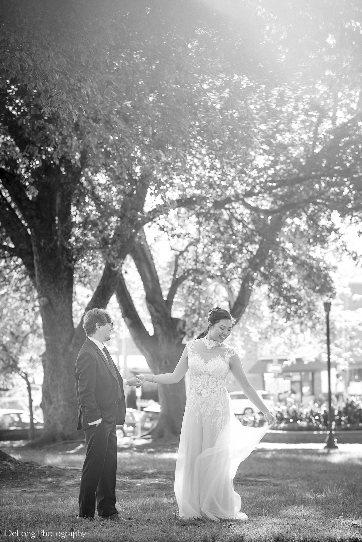 Bride and groom share a joyful moment under sun-dappled trees during their Wake County Courthouse elopement in downtown Raleigh, NC. The bride twirls in a sleeveless lace wedding jumpsuit, and the groom smiles in a dark suit. Captured by DeLong Photography.