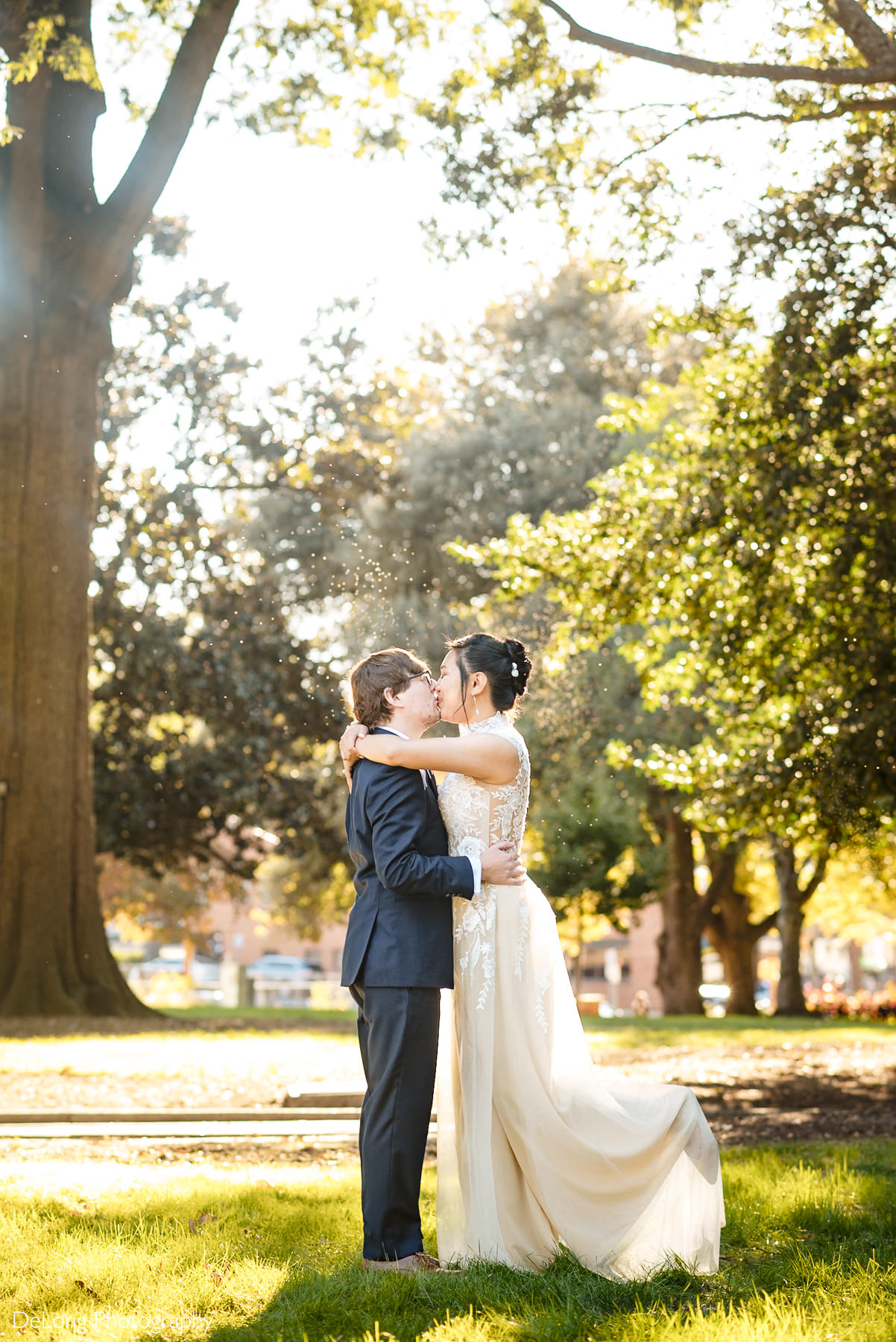 Bride and groom share a romantic kiss in the golden sunlight under the trees at Nash Square Park after their Wake County Courthouse elopement in Raleigh, NC. Captured by DeLong Photography.
