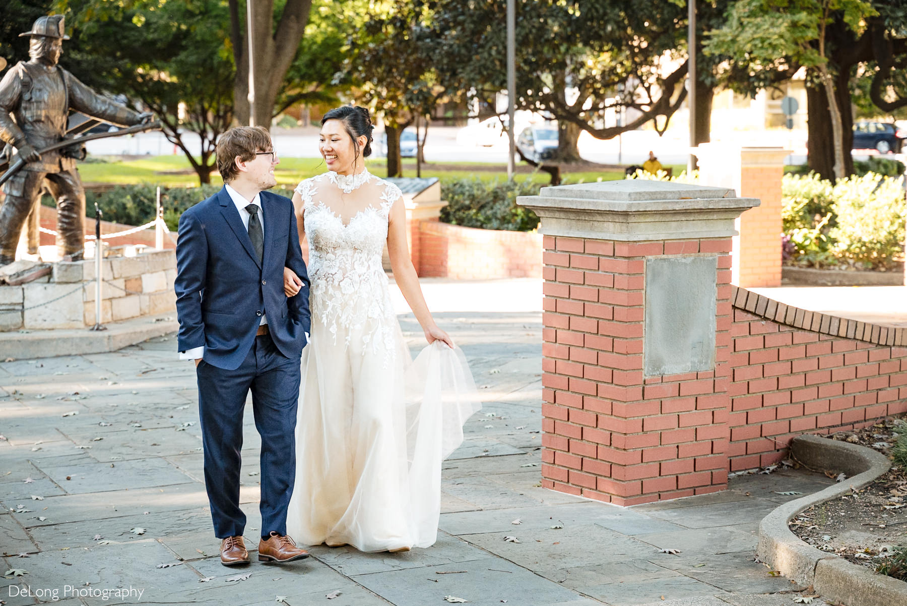 Bride and groom walk arm-in-arm, smiling joyfully during their Raleigh courthouse elopement at Nash Square. Captured by DeLong Photography, the bride wears a lace illusion wedding jumpsuit beside a firefighter statue.