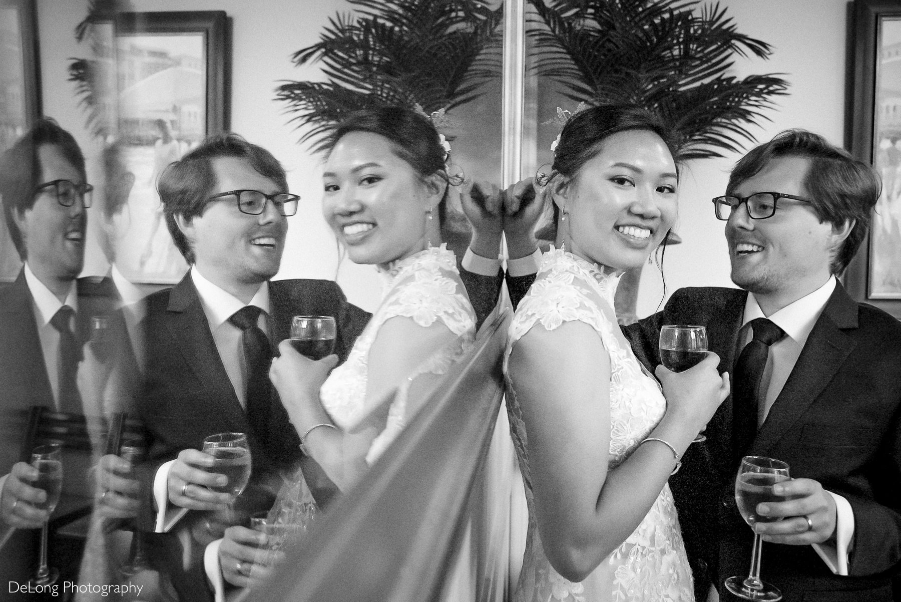 Bride and groom toast with wine glasses during their intimate Raleigh elopement reception, reflected in a mirror. The bride wears a lace high-neck wedding dress. The couple shares a joyful moment indoors at Caffe Luna after their Wake County ceremony. Captured by DeLong Photography.