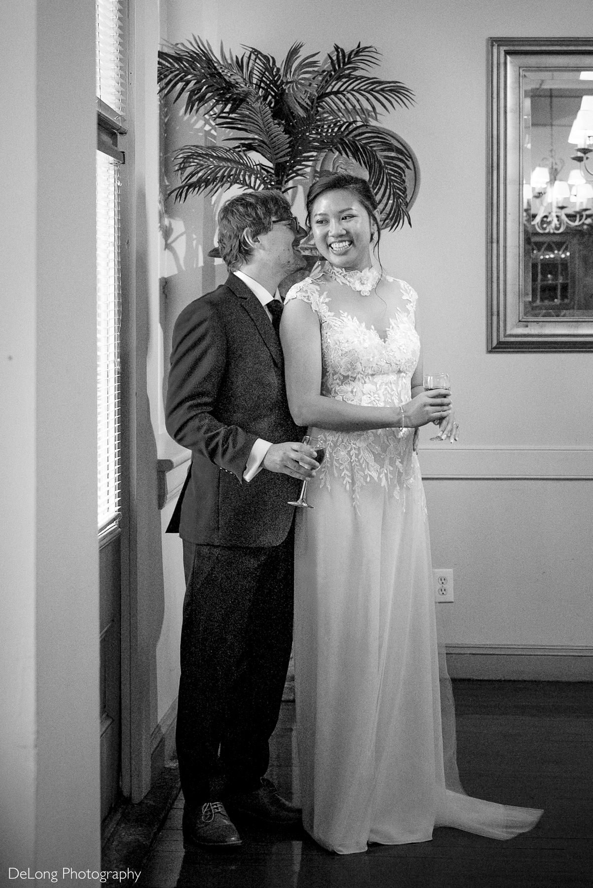 Bride and groom share a joyful moment at Caffe Luna with champagne glasses in hand during their Raleigh courthouse elopement reception. Captured by DeLong Photography, the bride wears a sleeveless lace wedding dress with illusion neckline.