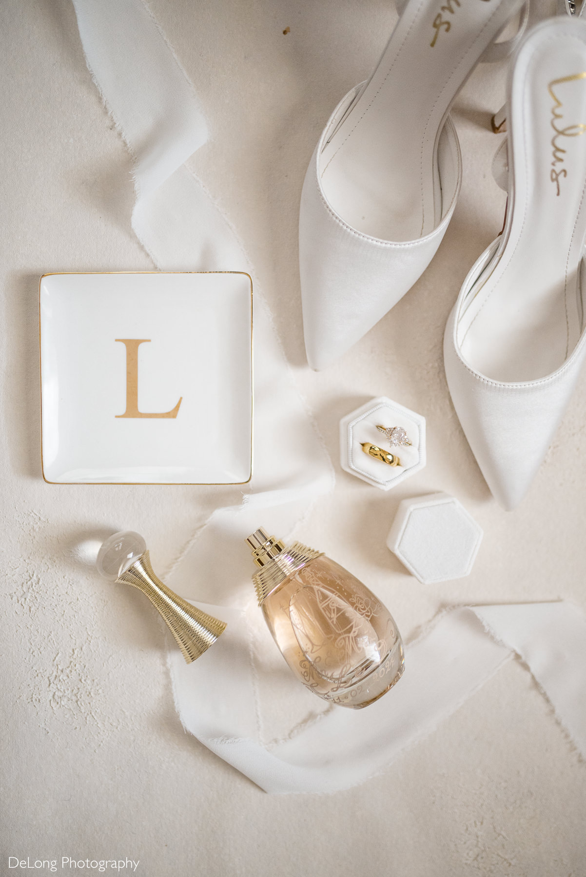 Flat lay of bridal details including white heels, wedding rings, perfume, and personalized jewelry dish, styled for a boho-glam wedding at The White Home in Rock Hill, SC. Photo by DeLong Photography.