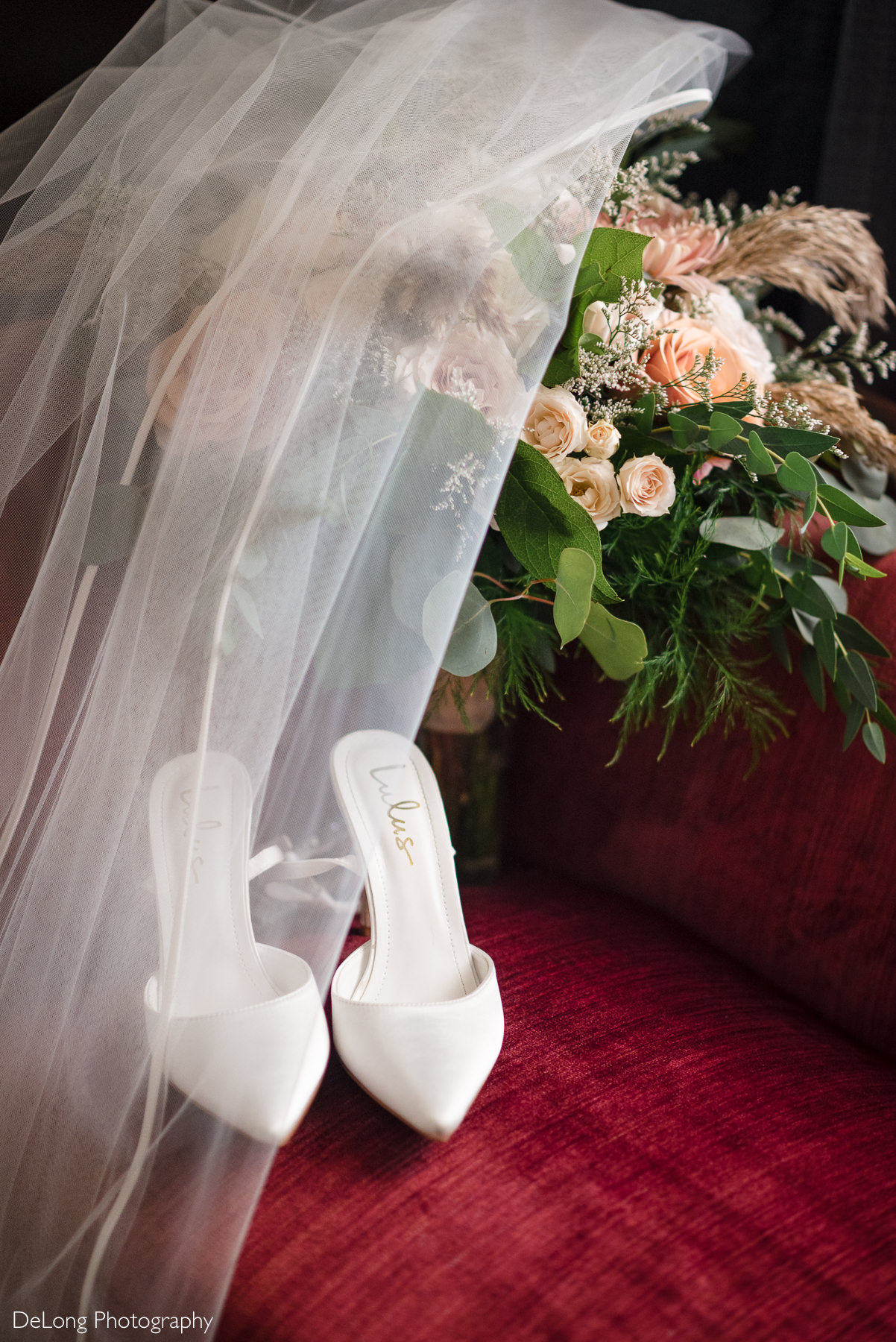 Wedding detail photo at The White Home in Rock Hill, SC. White bridal heels from Lulus rest on a red velvet chair with a flowing veil and pastel rose bouquet. Captured by a wedding photographer at The White Home specializing in elegant, romantic wedding photography.