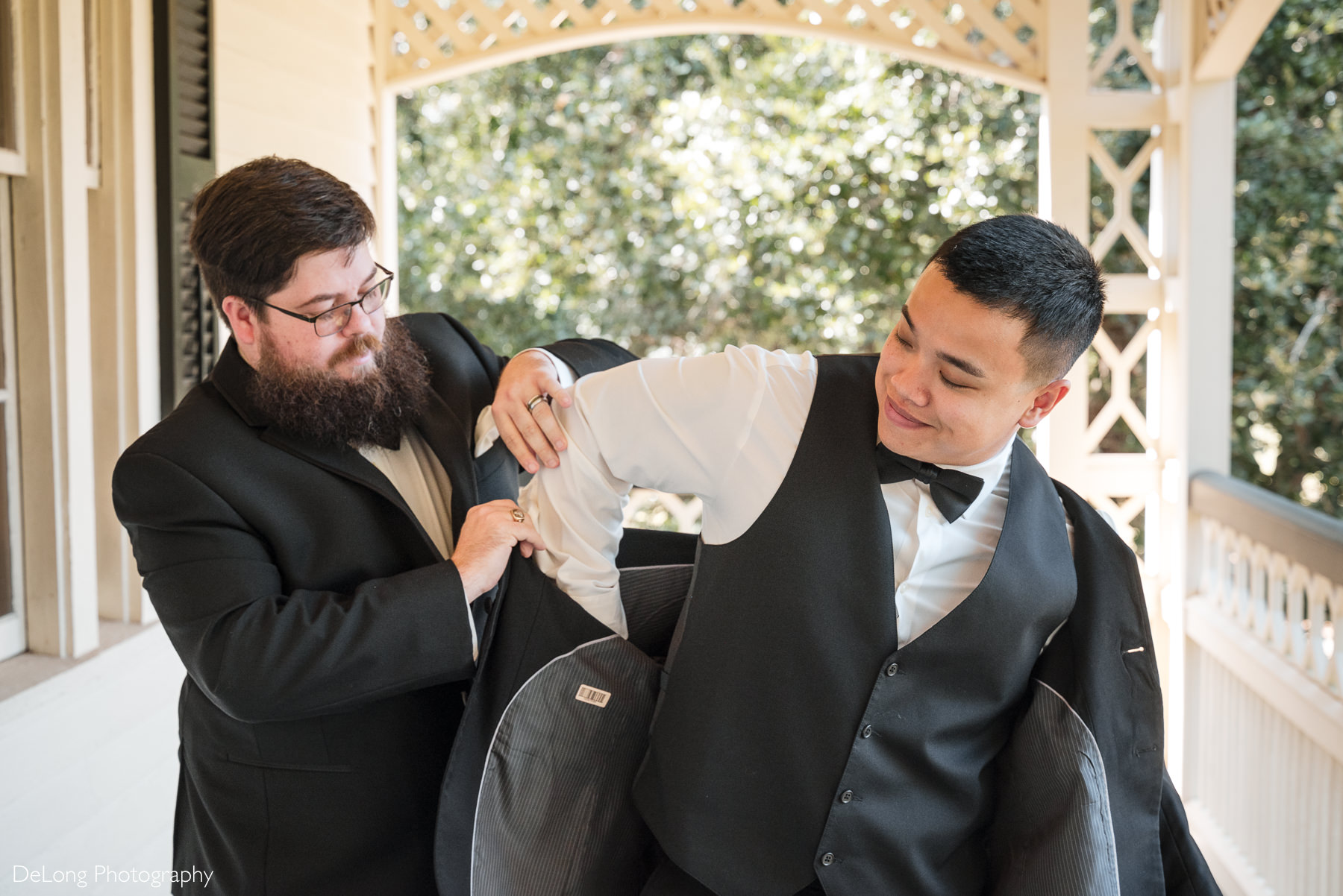 Groom getting ready at The White Home in Rock Hill, SC. A groomsman helps him into his black suit jacket on the historic porch before the wedding ceremony. Captured by a Rock Hill wedding photographer specializing in candid, natural wedding day moments.