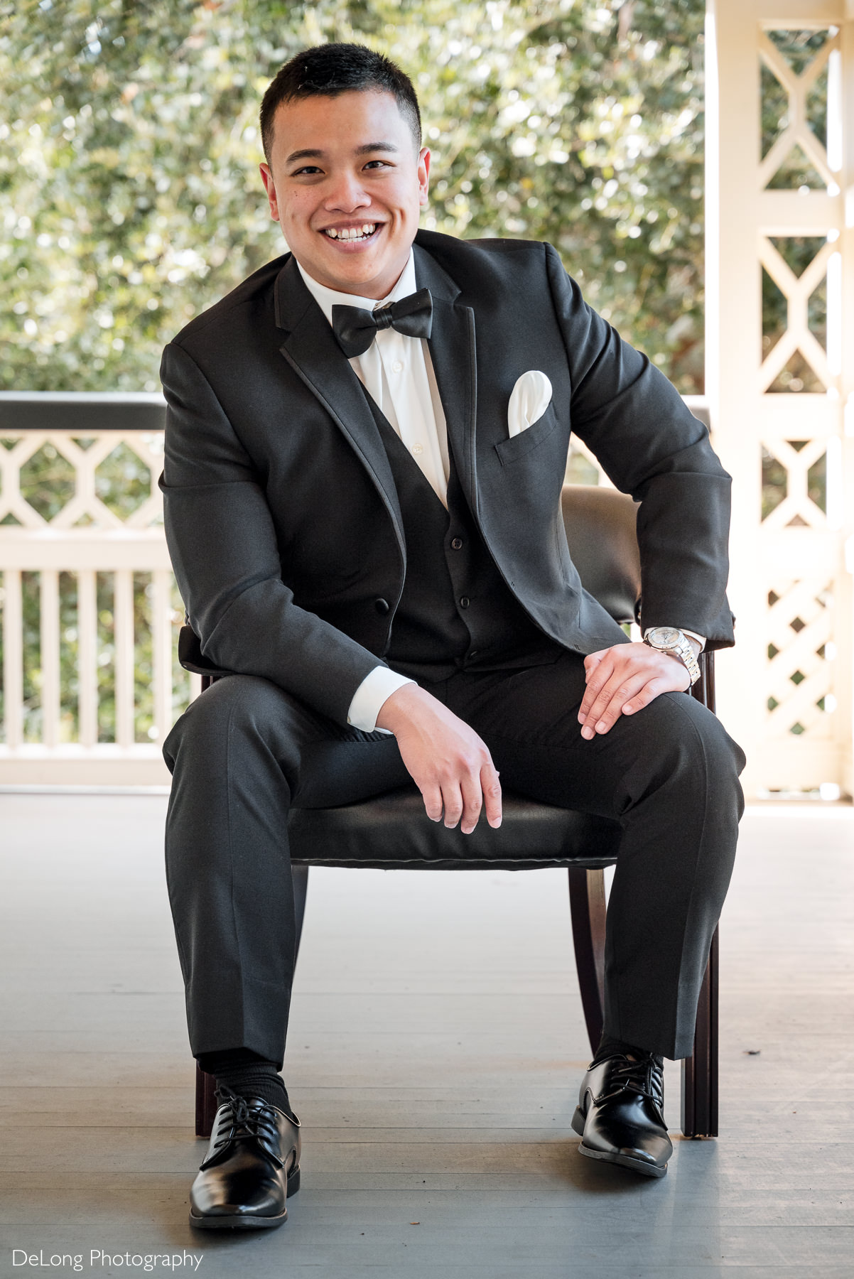 Groom portrait at The White Home wedding venue in Rock Hill, SC. Captured by DeLong Photography, this image shows the groom looking sharp in a black tuxedo with a bow tie and pocket square while seated on the wraparound porch. Perfect inspiration for couples searching for a Rock Hill wedding photographer at The White Home.