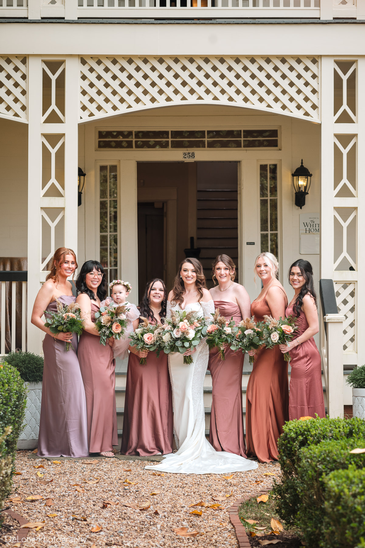 Bride with bridesmaids at The White Home wedding venue in Rock Hill, SC. The group poses in front of the historic home’s porch, holding bouquets of roses and greenery, showcasing elegant bridesmaid dresses in shades of mauve, blush, and rust. Perfect inspiration for bridal party portraits at The White Home Rock Hill wedding venue.