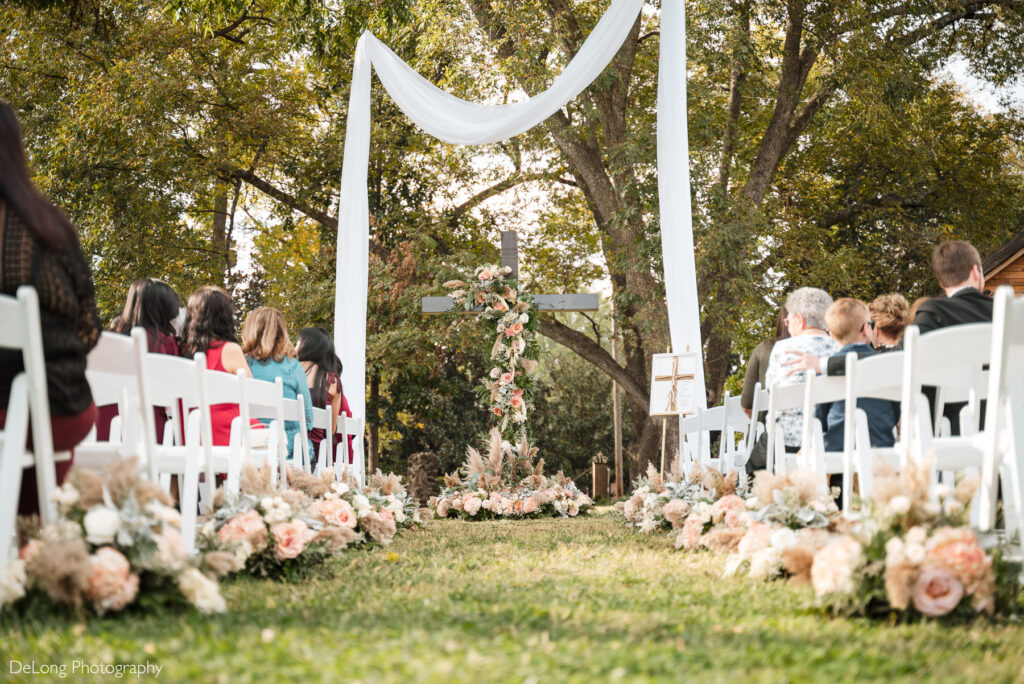 Outdoor wedding ceremony setup at The White Home in Rock Hill, SC. White chairs line both sides of the aisle decorated with lush blush and ivory floral arrangements. At the altar, a wooden cross adorned with cascading roses and pampas grass stands beneath flowing white drapery, framed by tall trees. Guests are seated, awaiting the bride and groom for a romantic garden wedding.