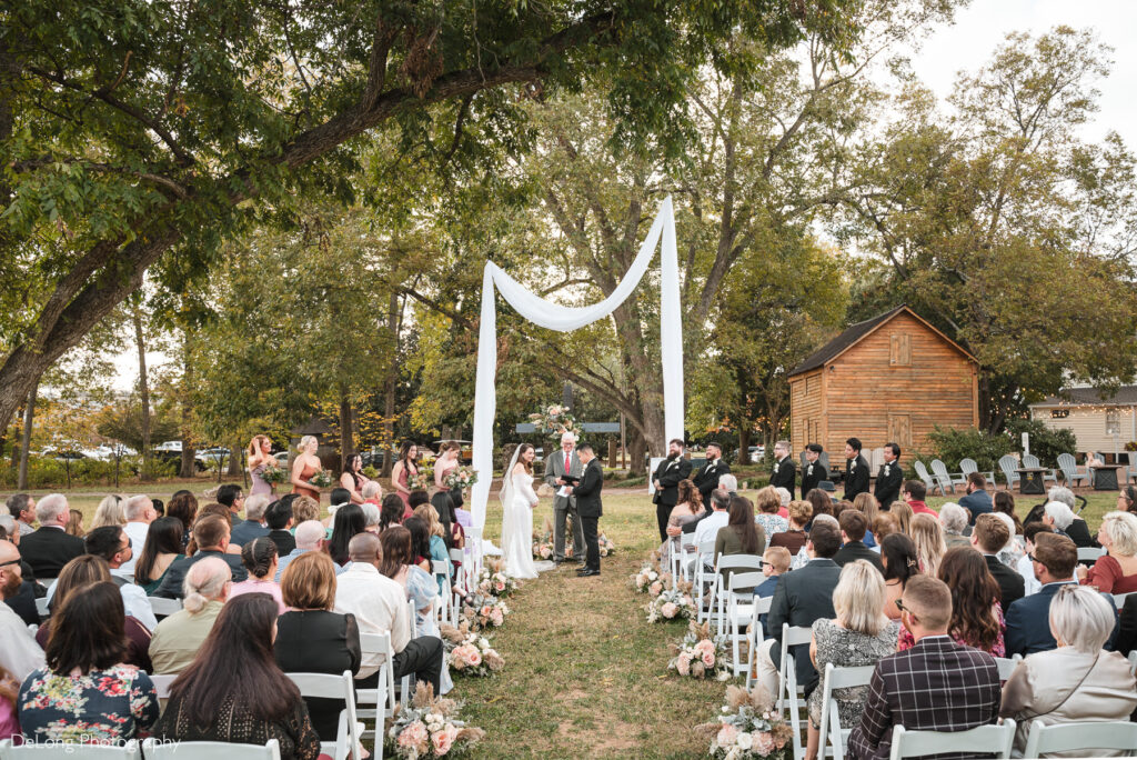 Fall outdoor wedding ceremony at The White Home in Rock Hill, SC with bride and groom exchanging vows under a white draped arch, surrounded by bridesmaids, groomsmen, and guests seated in rows decorated with blush floral arrangements.
