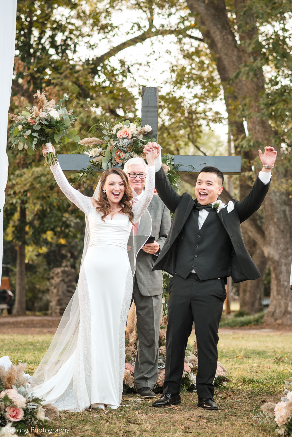 Bride and groom celebrating their wedding ceremony at The White Home in Rock Hill, SC. The couple stands in front of a floral cross with hands raised in joy, surrounded by lush greenery and romantic décor. Perfect example of a beautiful outdoor wedding venue in Rock Hill, South Carolina.