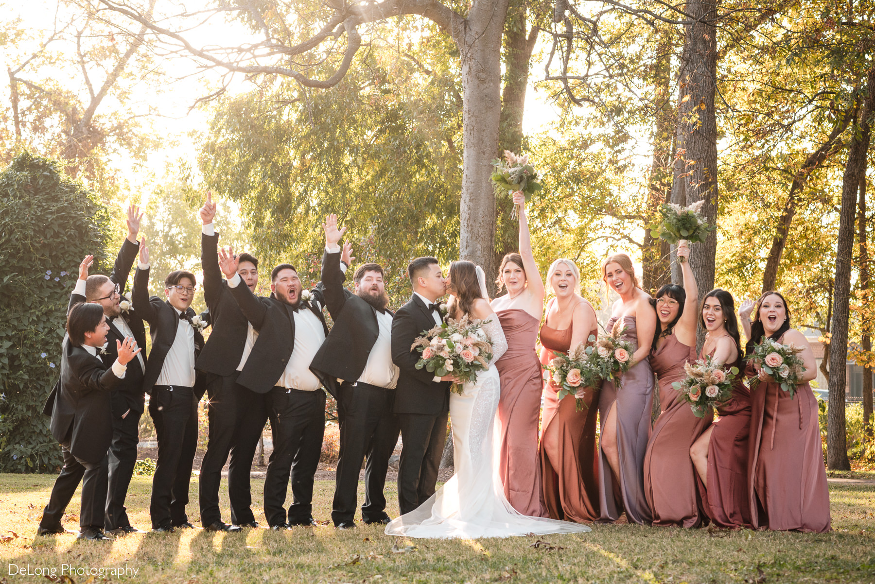 Wedding party photo at The White Home in Rock Hill, SC, with the bride and groom sharing a kiss while surrounded by cheering bridesmaids in mauve and rust-colored dresses and groomsmen in black tuxedos. Captured in golden sunlight, this joyful bridal party portrait highlights the vibrant fall wedding atmosphere at The White Home, a popular Rock Hill wedding venue.
