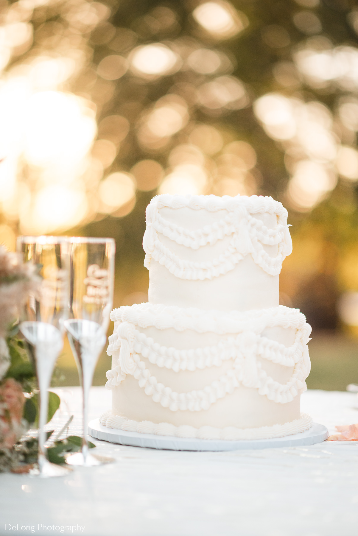 Classic two-tier white wedding cake decorated with piped buttercream swags, displayed outdoors at The White Home in Rock Hill with champagne flutes beside it.