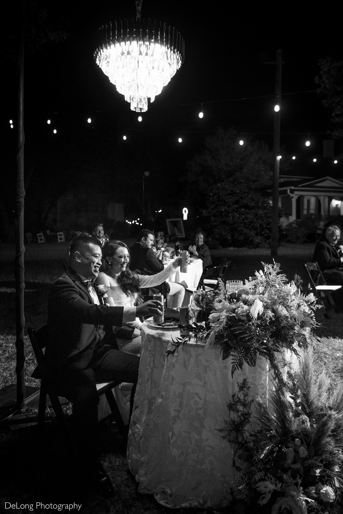 Bride and groom smiling and raising drinks together at their sweetheart table under a chandelier and string lights during their outdoor wedding reception at The White Home in Rock Hill.