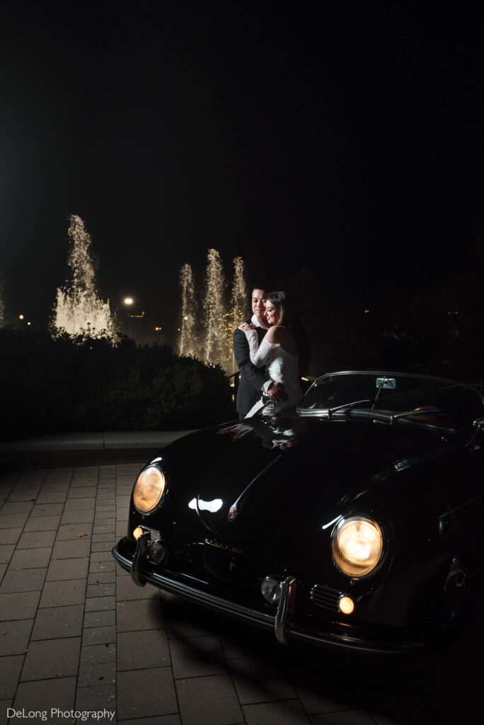 Bride and groom embrace by a vintage black Porsche with glowing headlights and illuminated fountains in the background during their Rock Hill wedding.