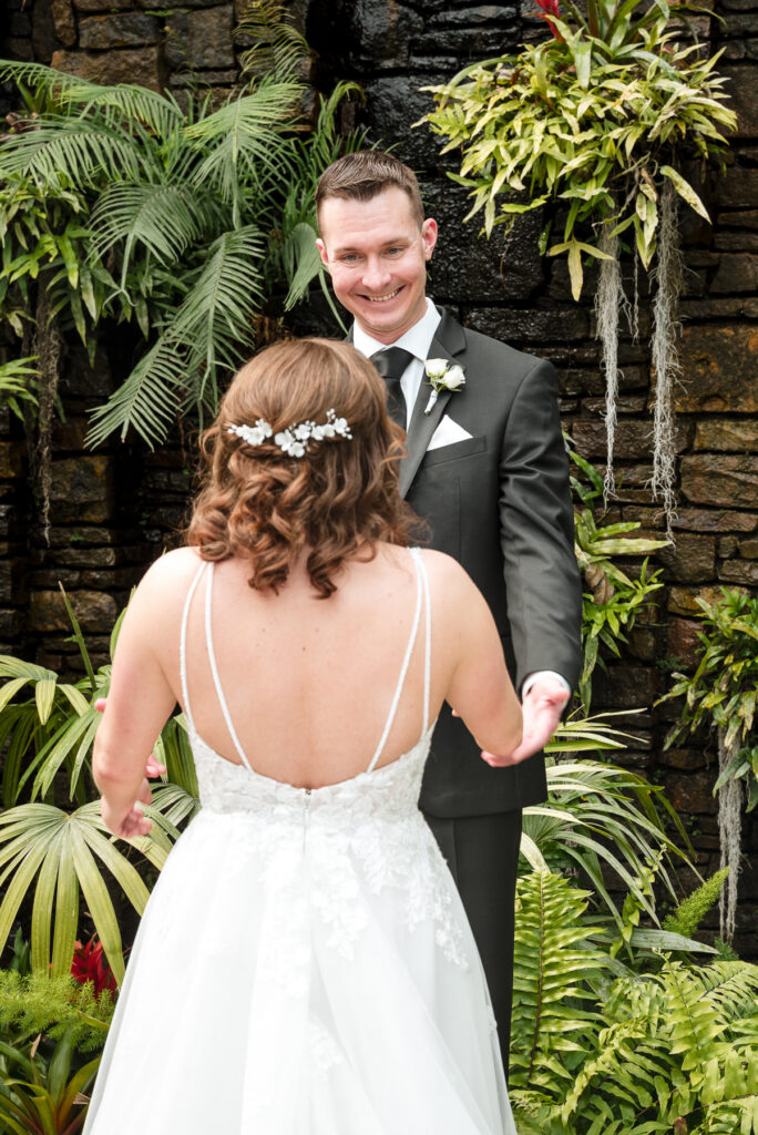 Groom smiles as he sees his bride during a first look at a spring wedding at Daniel Stowe Botanical Garden in Belmont, NC, surrounded by lush greenery and tropical plants.