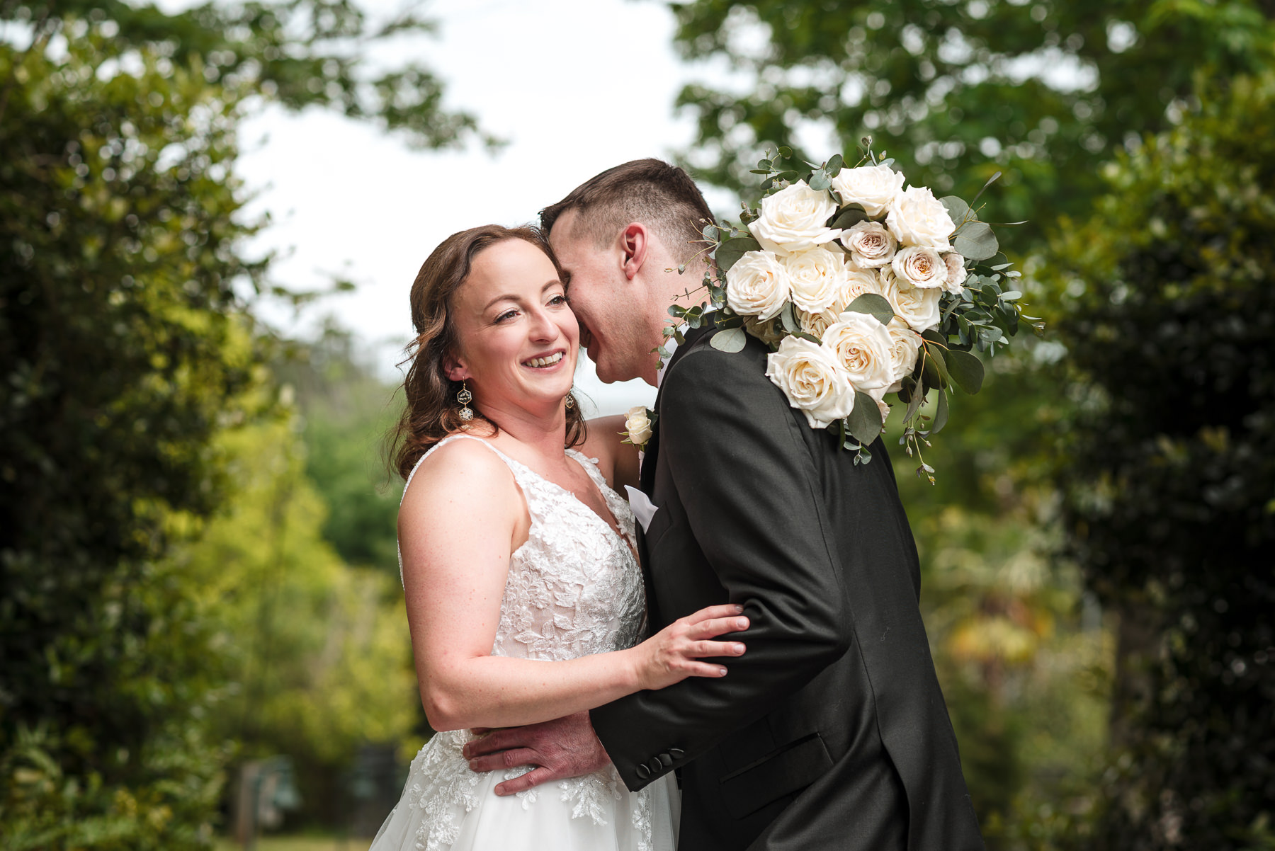 Bride and groom embrace beneath lush greenery during a spring wedding at Daniel Stowe Botanical Garden in Belmont, NC, with the bride’s bouquet of white roses and eucalyptus draped over the groom’s shoulder. Romantic outdoor wedding photography by DeLong Photography captures the joyful couple in a scenic garden setting near Charlotte, North Carolina.