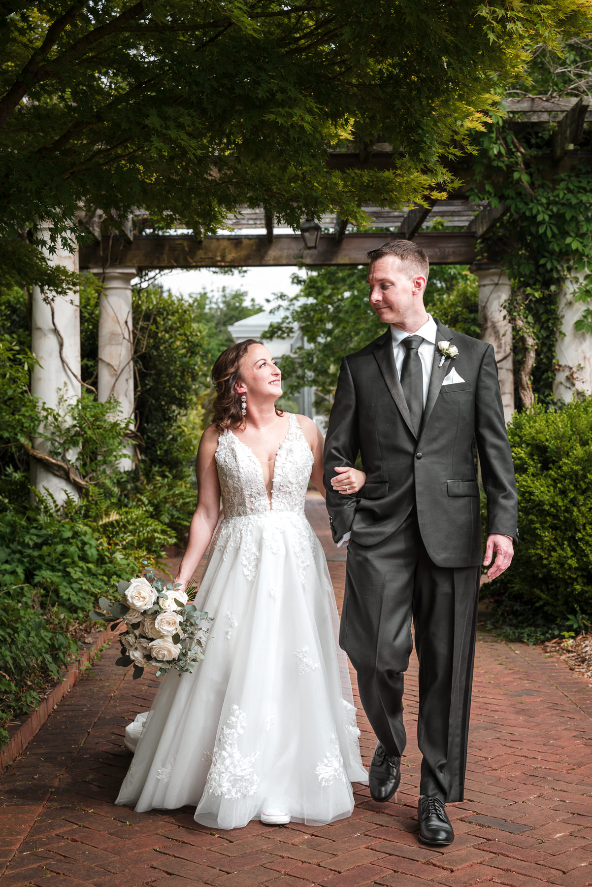 Bride and groom walk hand in hand beneath a garden arbor during a spring wedding at Daniel Stowe Botanical Garden in Belmont, NC, surrounded by lush greenery and brick pathways.