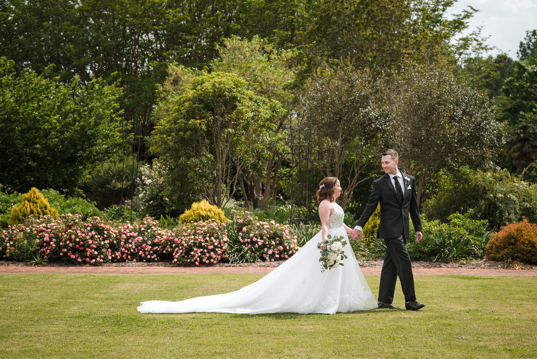 Bride and groom walk across the lawn during a spring wedding at Daniel Stowe Botanical Garden in Belmont, NC, with blooming pink flowers and lush greenery in the background.