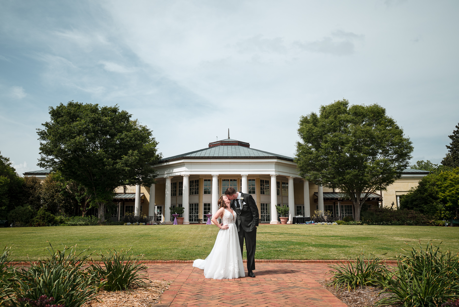 Bride and groom share a kiss on the lawn in front of the grand white columns at Daniel Stowe Botanical Garden in Belmont, NC during a spring wedding celebration. Captured by DeLong Photography near Charlotte, North Carolina, this timeless botanical garden wedding portrait highlights the elegant architecture and scenic outdoor setting.