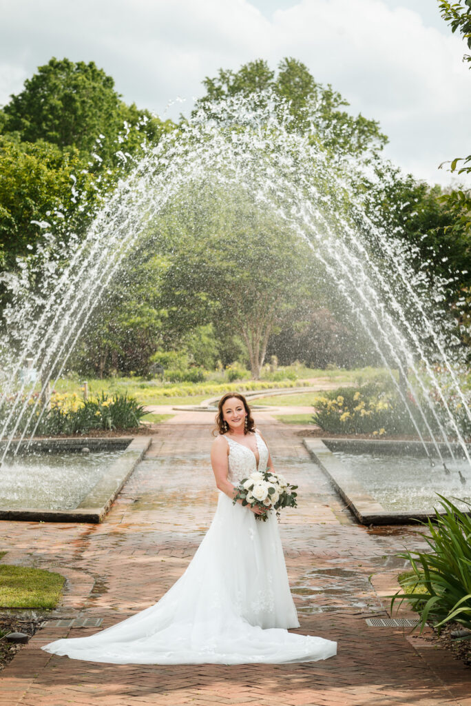 Bride standing beneath the fountain at Daniel Stowe Botanical Garden in Belmont, NC during a spring wedding, holding a white rose and greenery bouquet on a brick pathway surrounded by lush gardens and blooming flowers.