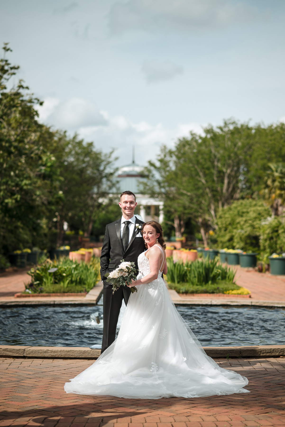 Bride and groom pose in front of a reflecting pool and manicured gardens at Daniel Stowe Botanical Garden in Belmont, NC during their spring wedding celebration. Captured by DeLong Photography near Charlotte, North Carolina, this elegant botanical garden wedding portrait highlights the fountain, lush greenery, and the bride’s flowing lace gown.