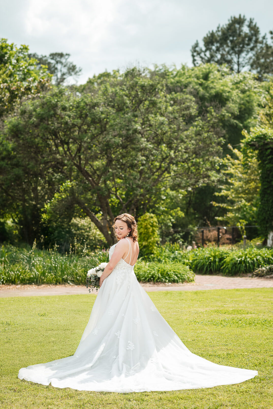 Bride in a flowing lace wedding gown holding a white and greenery bouquet during a spring wedding portrait at Daniel Stowe Botanical Garden in Belmont, North Carolina, surrounded by lush gardens and bright green lawn.